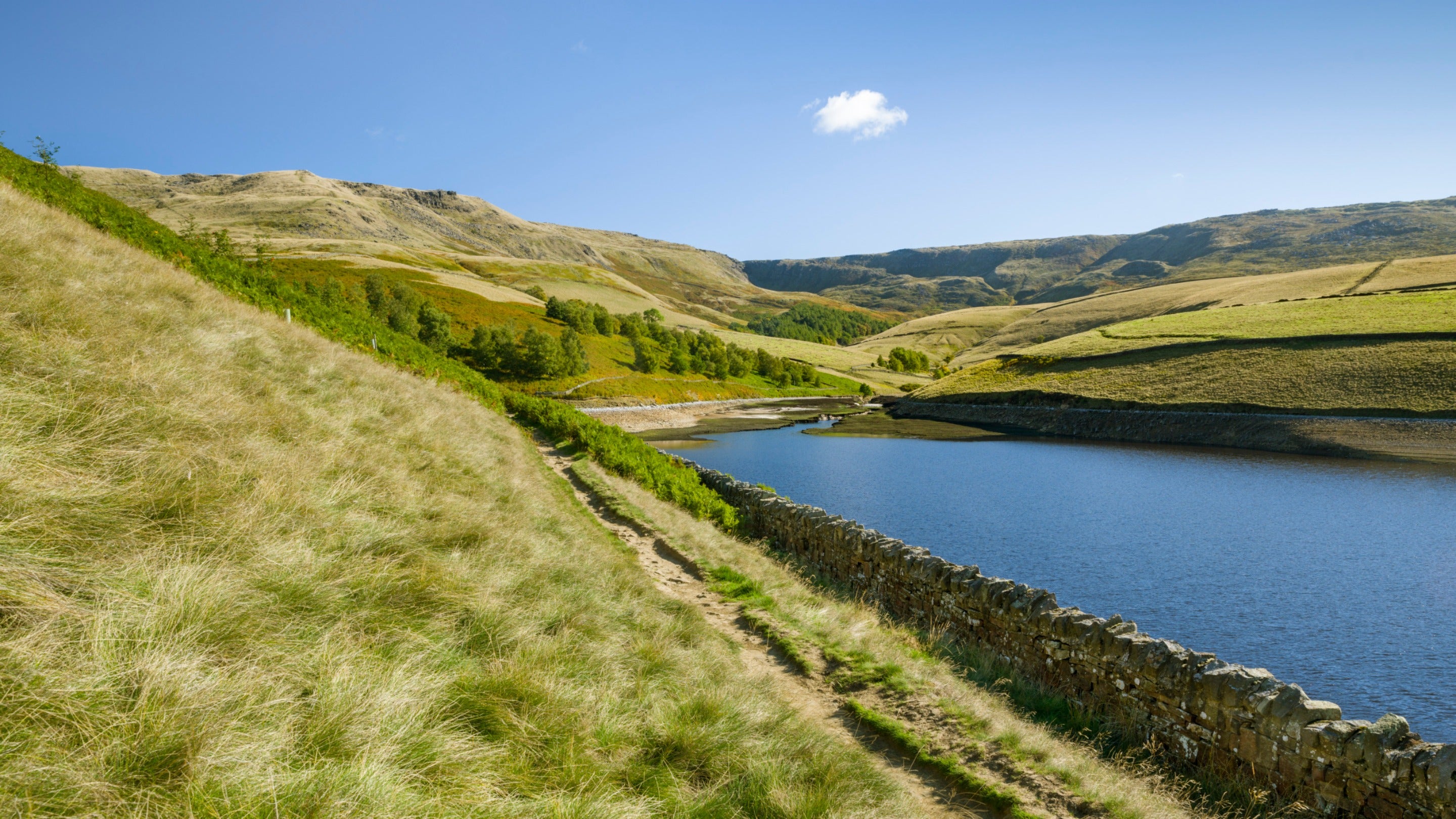 A view across Kinder Reservoir looking towards Kinder Scout on a sunny day, with a hill on the left side of the image and a stone wall surrounding the reservoir