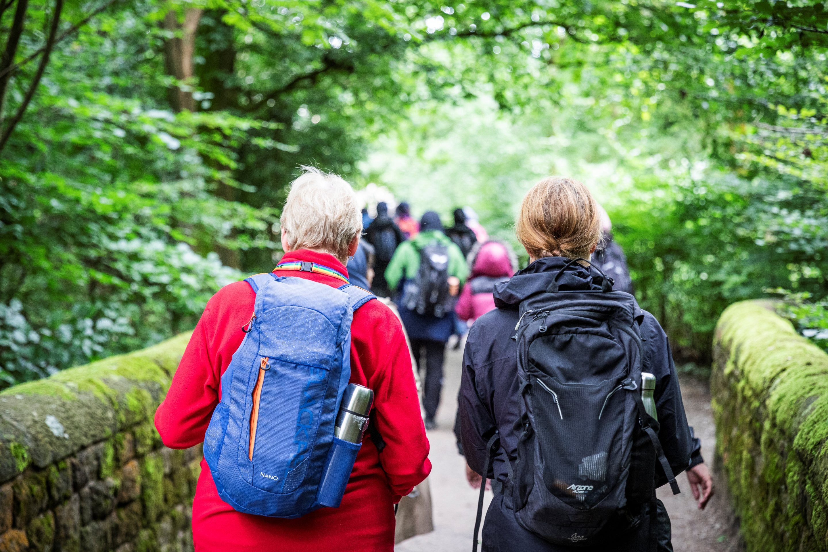 Two people walking together through woodland wearing  back packs. There is a group of people in front of them in the distance.