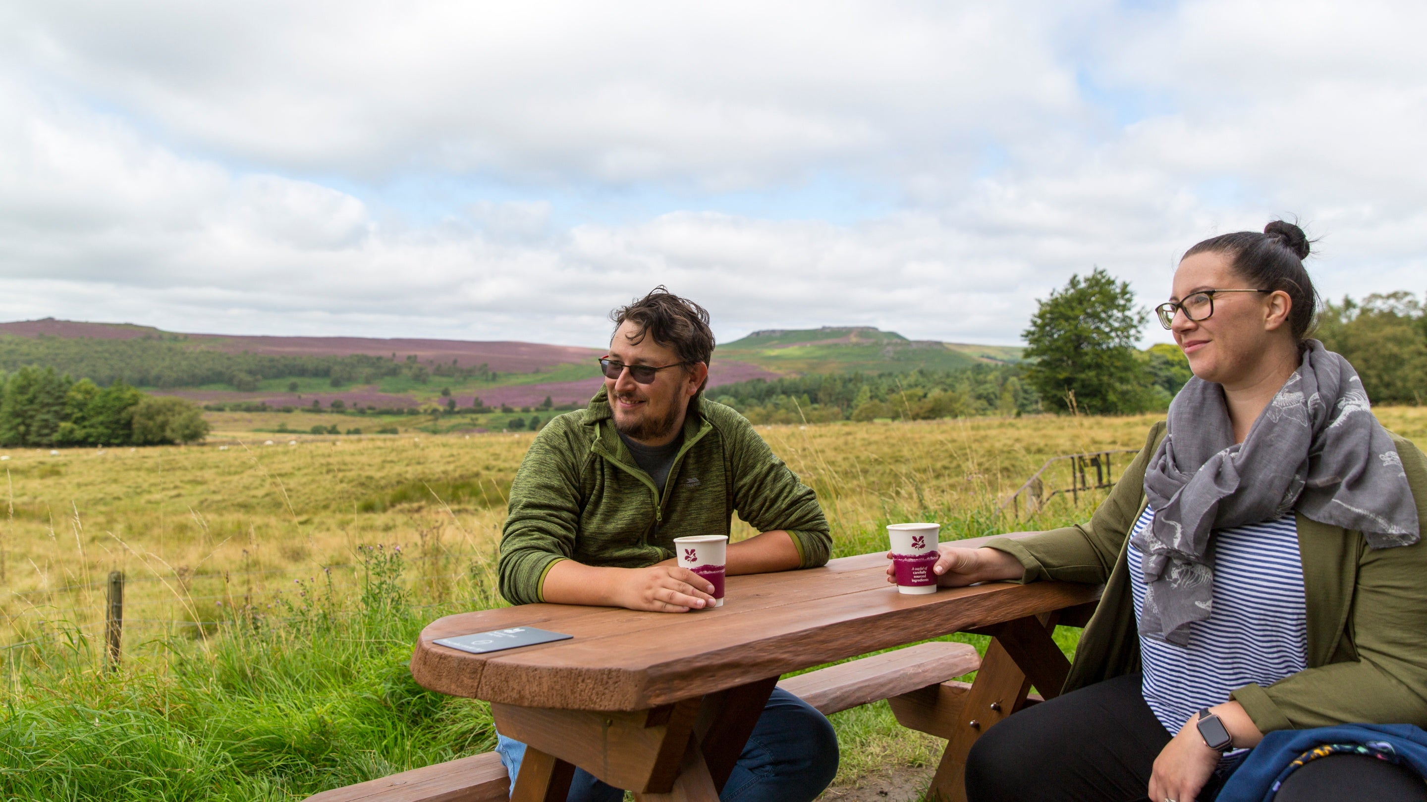 A couple sit at a picnic table drinking coffees with a country view behind them