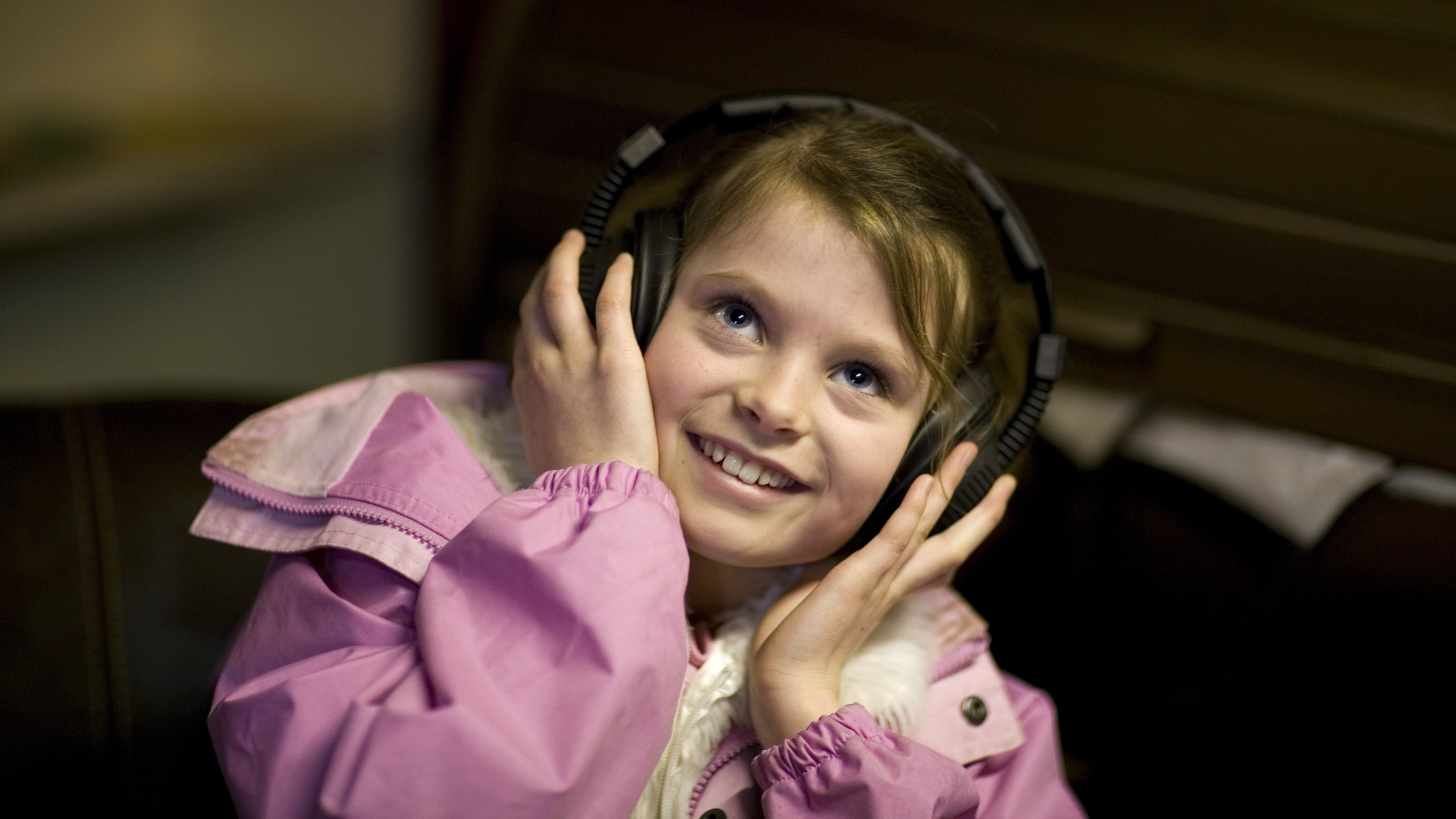 A child wears big black headphones and smiles at the camera in front of a dark background