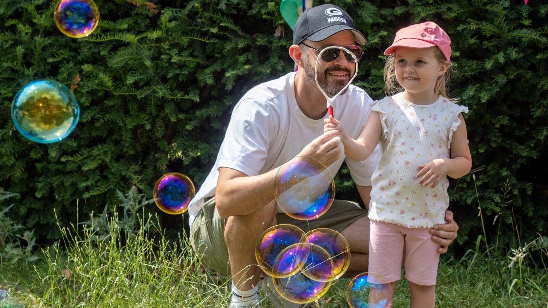 A child standing next to a crouching adult playing with bubbles. Both are in grass with a hedge in the background and surrounded by bubbles