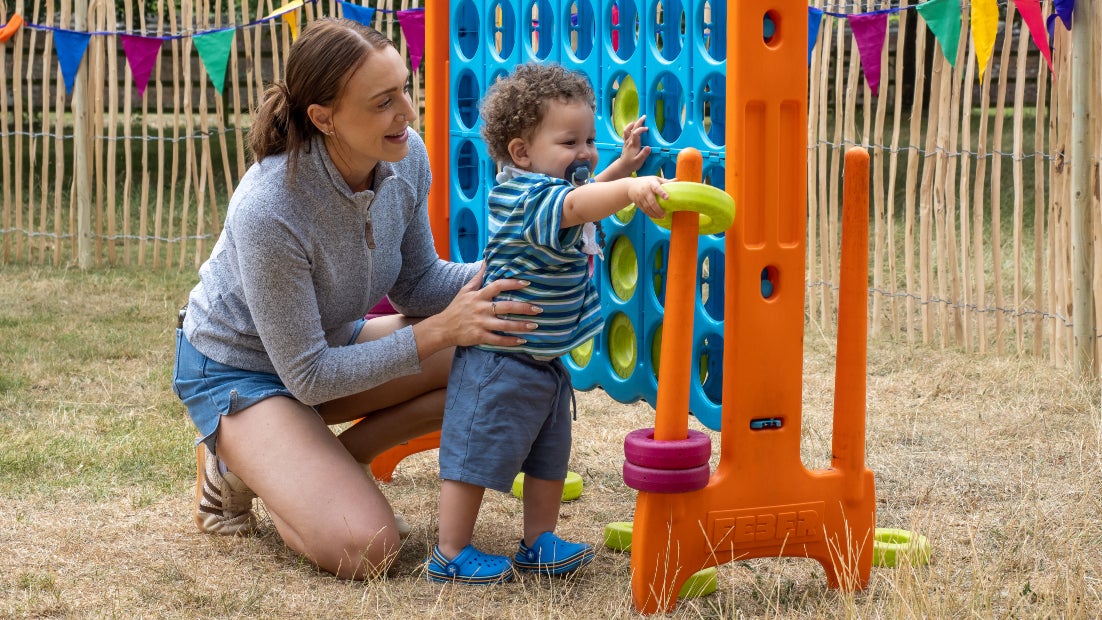 A child standing next to a crouching adult playing with a giant connect 4 game in a grass activities area at Summer of Play