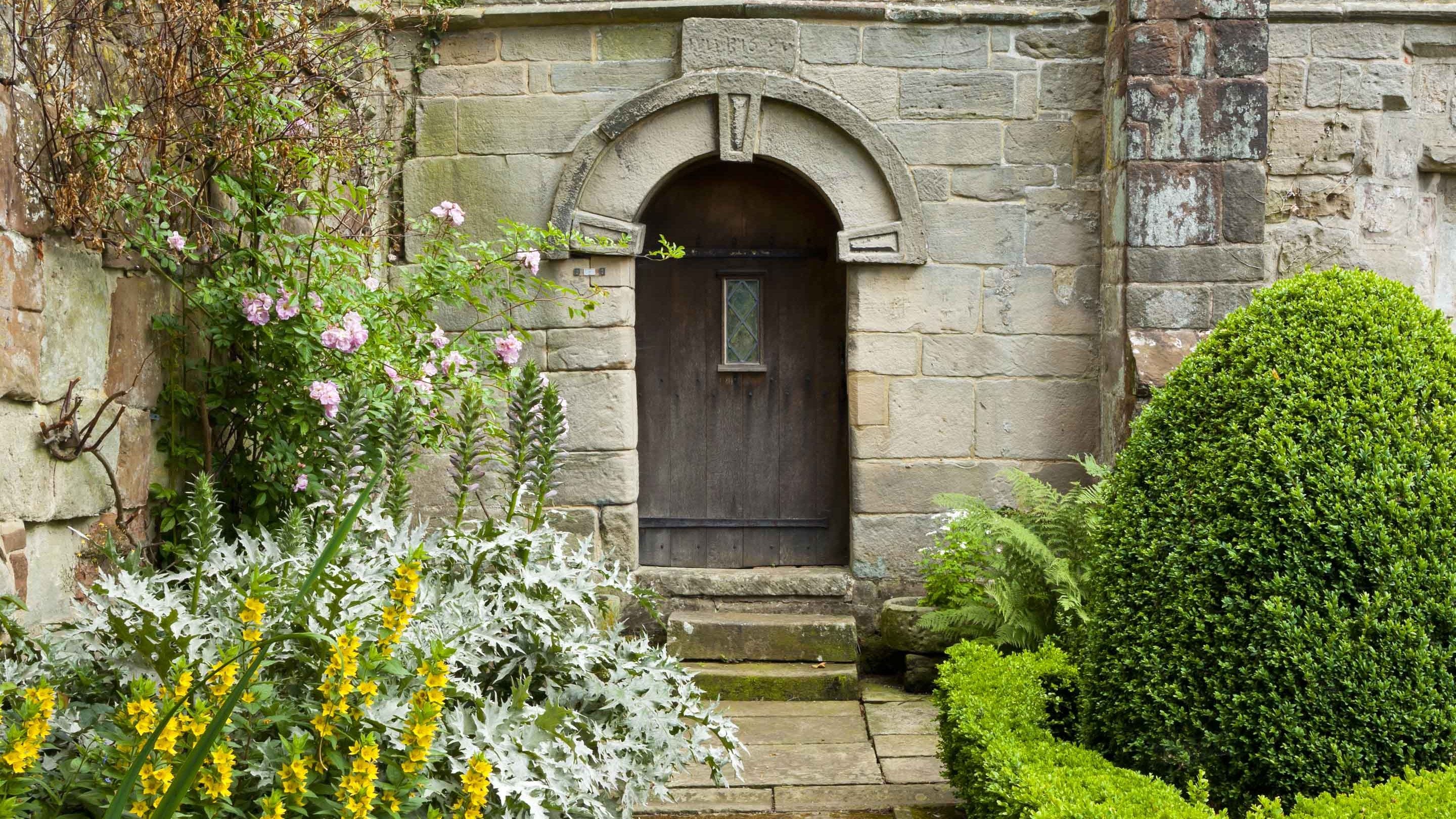 View across the herb knot garden, with a stone path and a round-arched wooden door in a stone wall, towards the medieval range of The Old Manor, Norbury, Derbyshire.