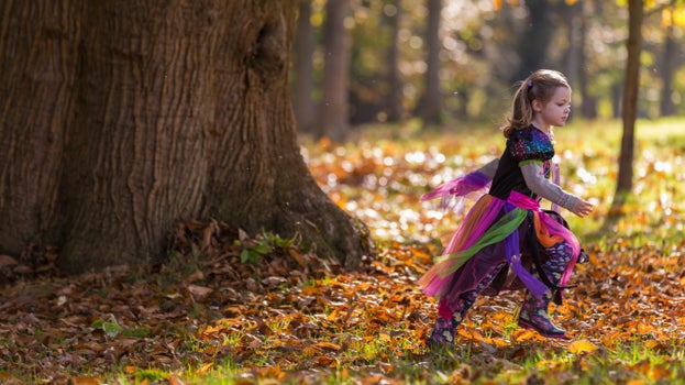 Young child wearing a bright fairy costume with colourful ribbons and patterned boots runs across a carpet of golden-brown autumn leaves beside a large tree trunk in Petworth House and Park, West Sussex, with sunlight filtering through the trees in the background.