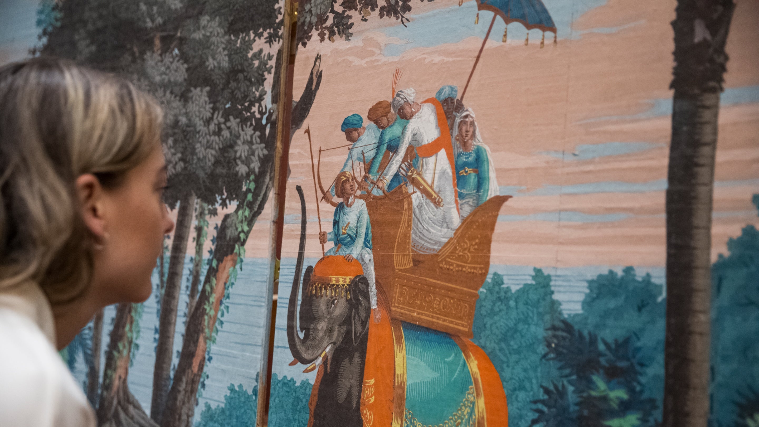 A visitor looks closely at a large, detailed mural showing a richly decorated elephant carrying several figures in traditional clothing. The scene is set against a backdrop of tall trees, foliage, and a soft pastel sky.