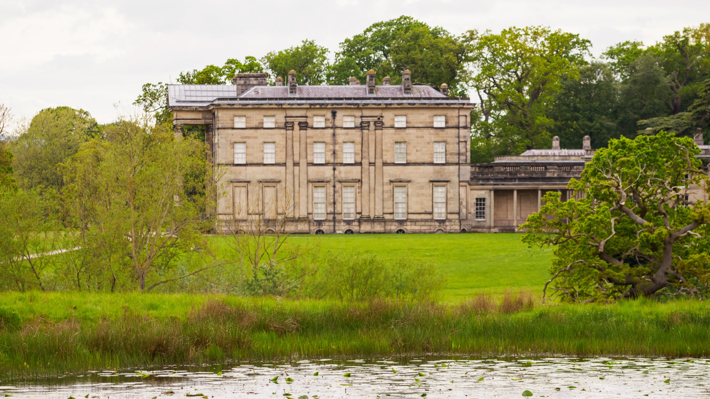 A large historic stone mansion set within expansive green parkland, viewed from across a pond with reeds in the foreground. The building has tall sash windows, a symmetrical façade and is surrounded by mature trees under a lightly cloudy sky
