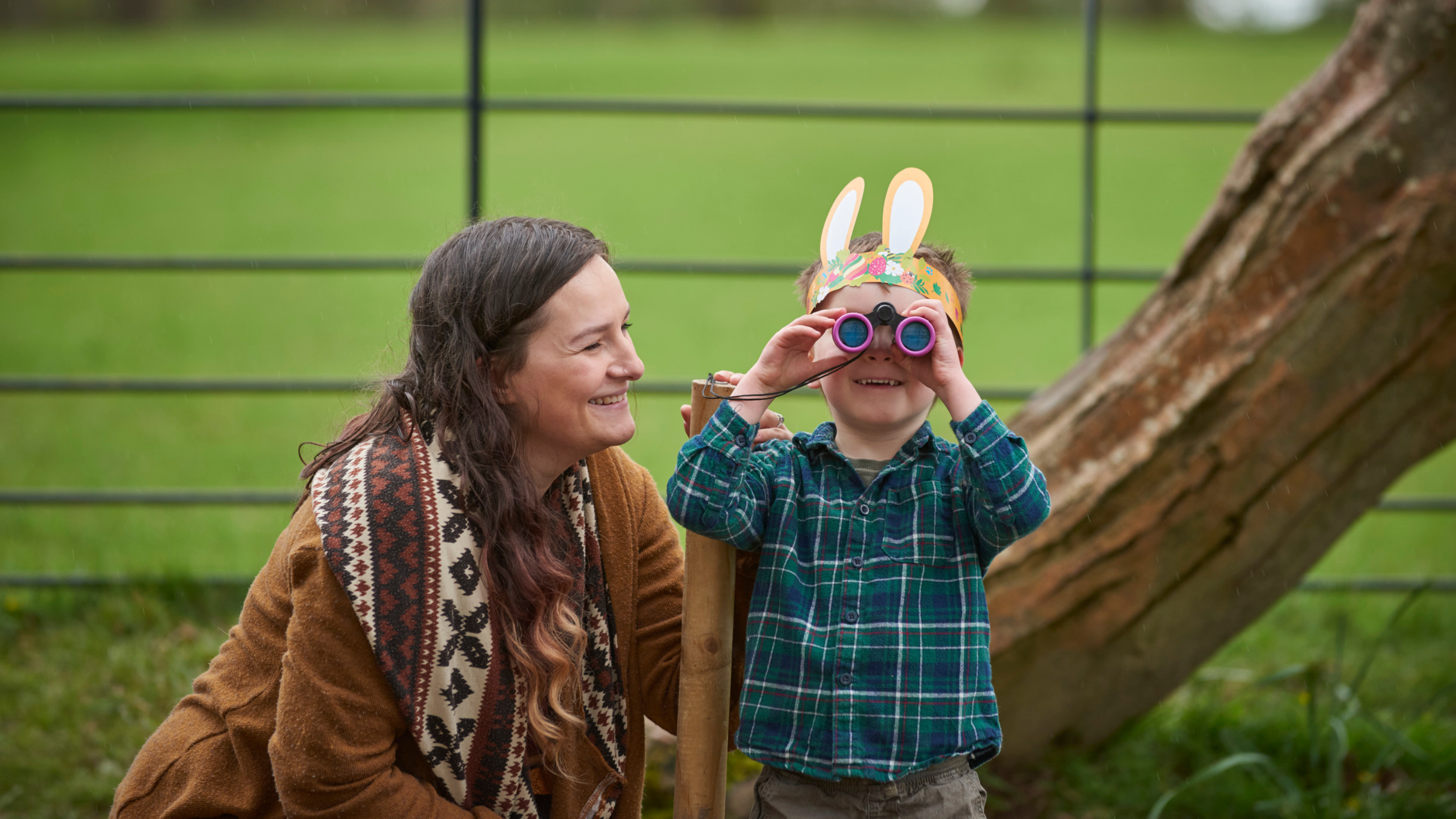 A woman and a young child are outdoors in a green park setting. The woman, wearing a brown coat and patterned scarf, smiles warmly at the child. The child, dressed in a green plaid shirt, wears a handmade bunny ear headband and looks through a pair of colourful binoculars. They appear to be engaging in a fun outdoor activity together. A wooden post, a tree trunk, and a metal fence are visible in the background.