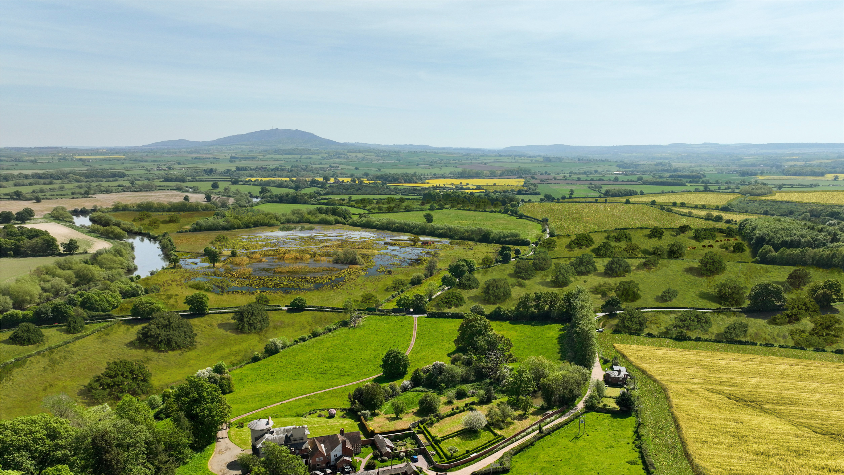 An aerial view of a picturesque rural landscape featuring a mix of lush green fields, a meandering river, a wetland area, and scattered trees.