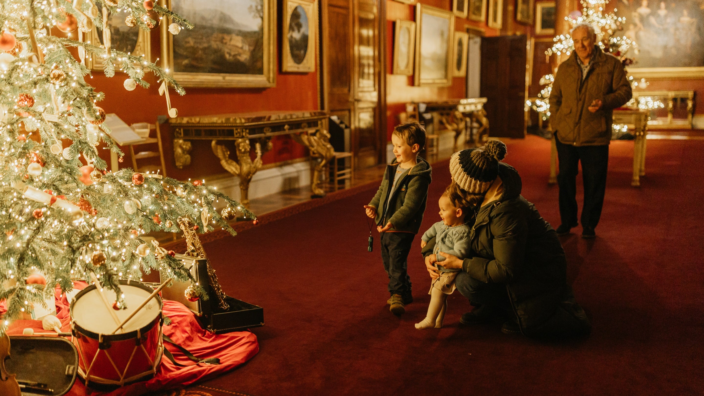 A family walks through a festively room at Attingham Park in Shropshire during Christmas. The scene features twinkling fairy lights strung across trees, historic architecture illuminated in warm tones, and children pointing excitedly at seasonal displays, evoking a joyful holiday atmosphere.
