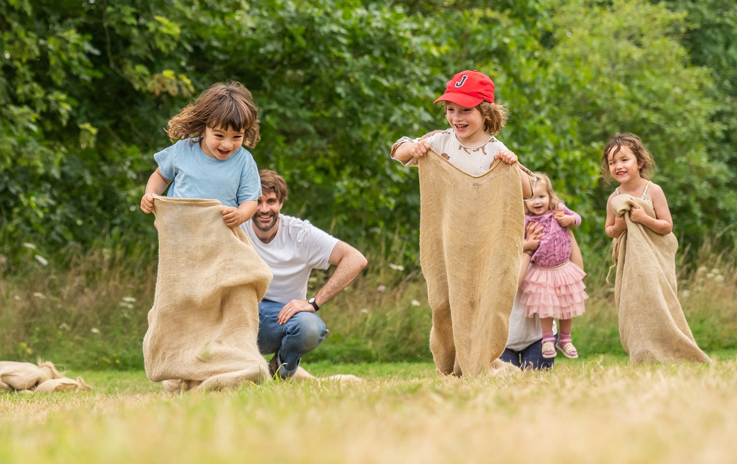 Three children in hessian sacks racing each other as two adults watch.