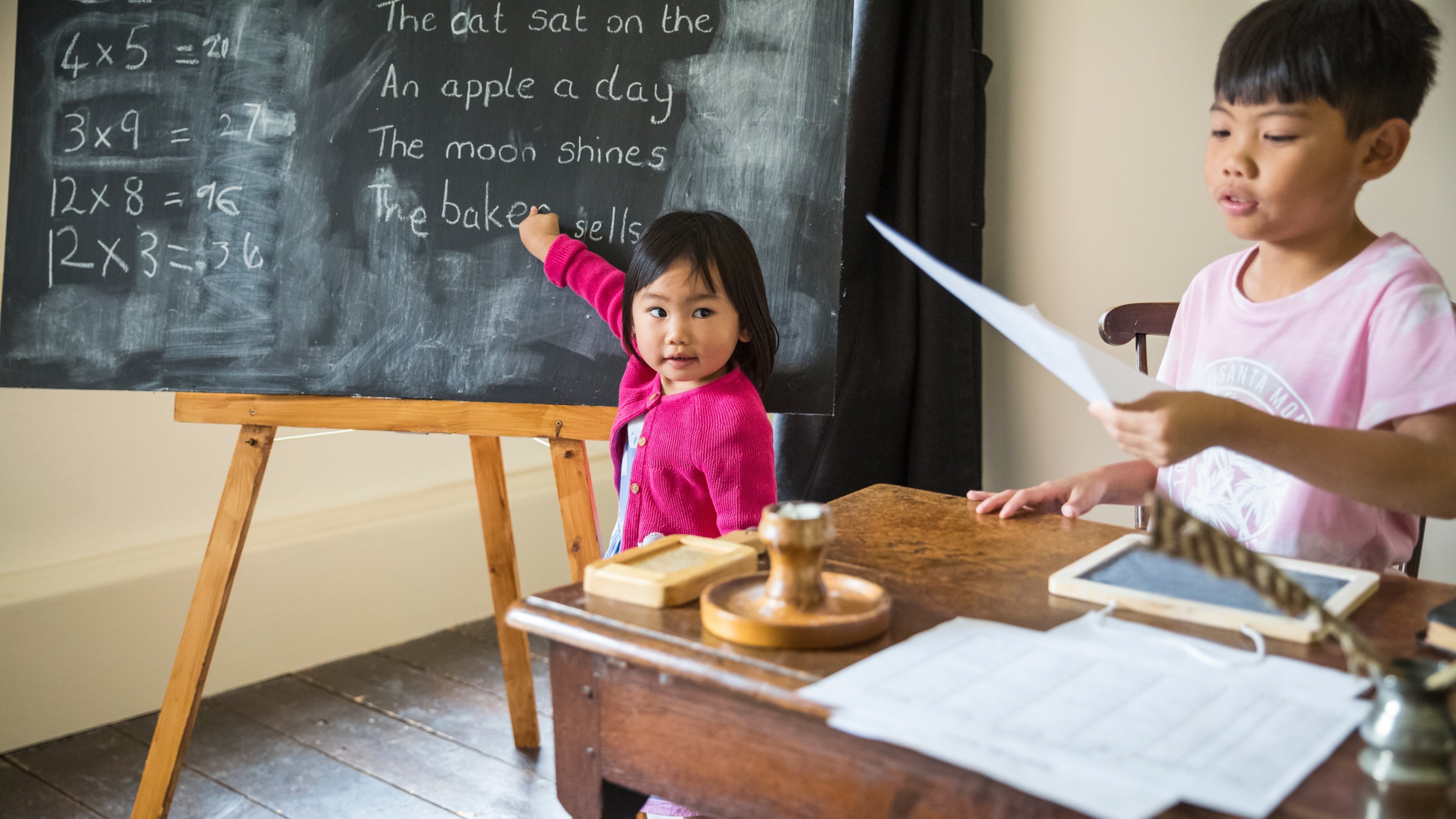 A young family are looking through papers at an old school desk with a quill on top, while a young girl in a pink cardigan is pointing at a blackboard.