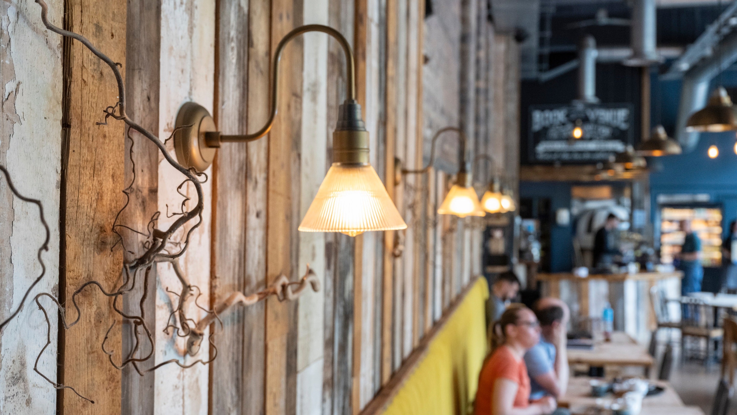 Close up of a Victorian style light fitting on a wall fronted with wood. In the background diners sit at a table inside the Furnace Kitchen restaurant