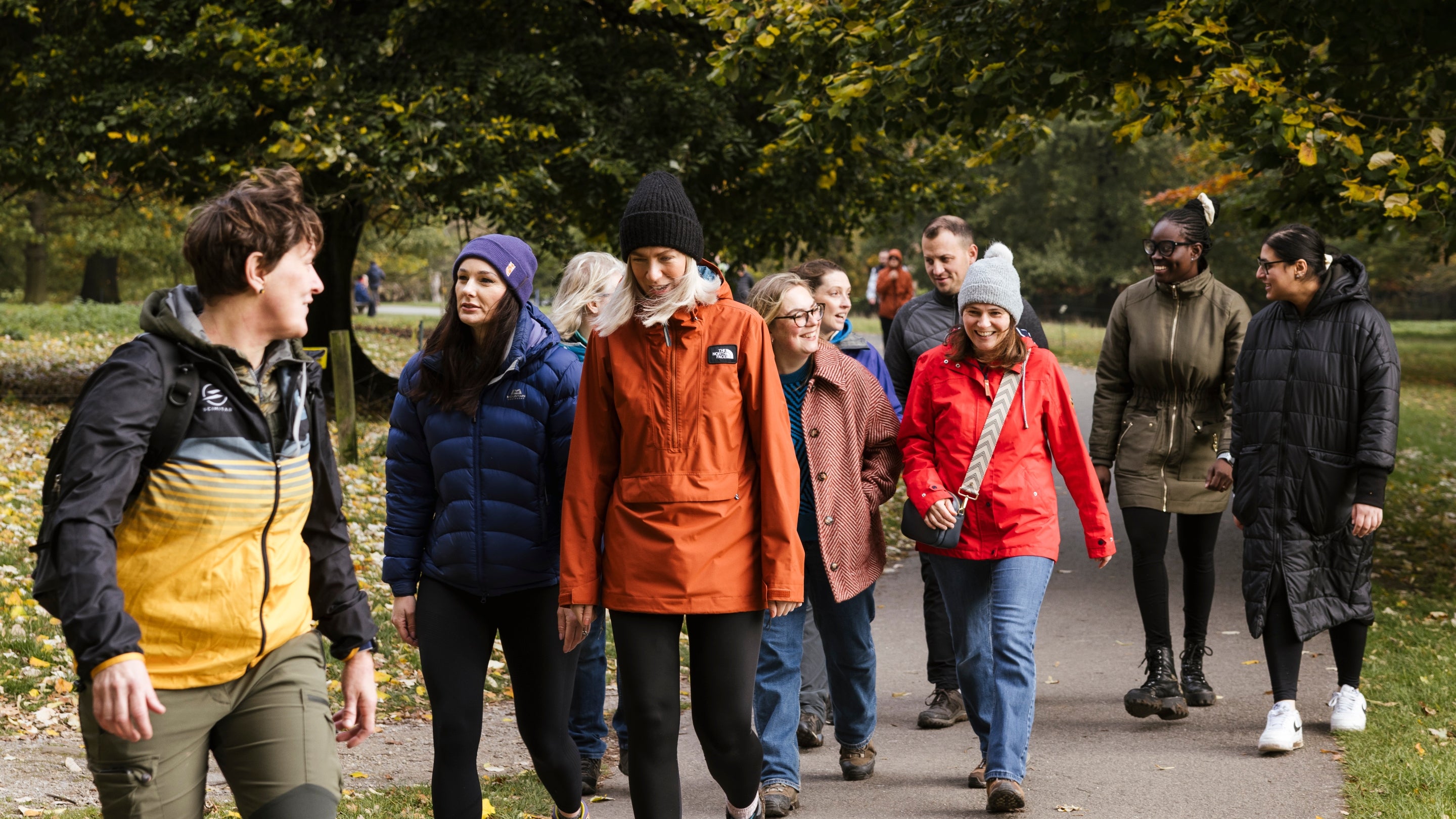 Group of visitors with happy faces, chatting and enjoying a walk with slightly autumnal leaves on large trees in the background.