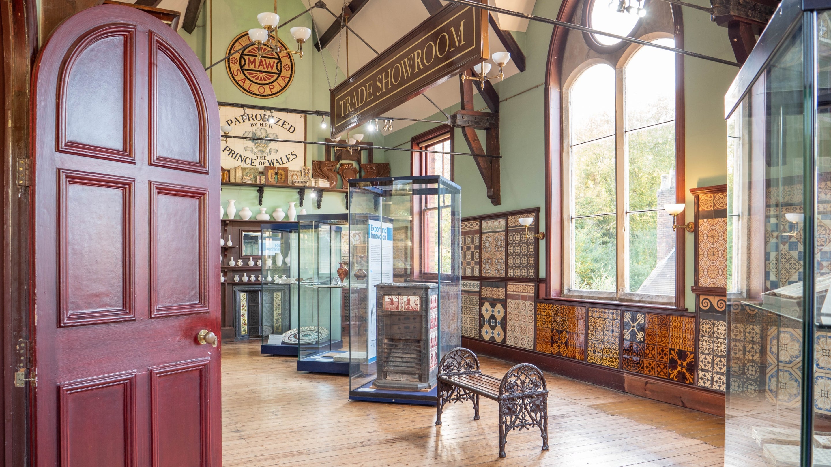 A view looking through a dark red door into a brightly lit museum room with ornate windows and a wooden floor. Around the room are examples of different tiles with beautiful designs.