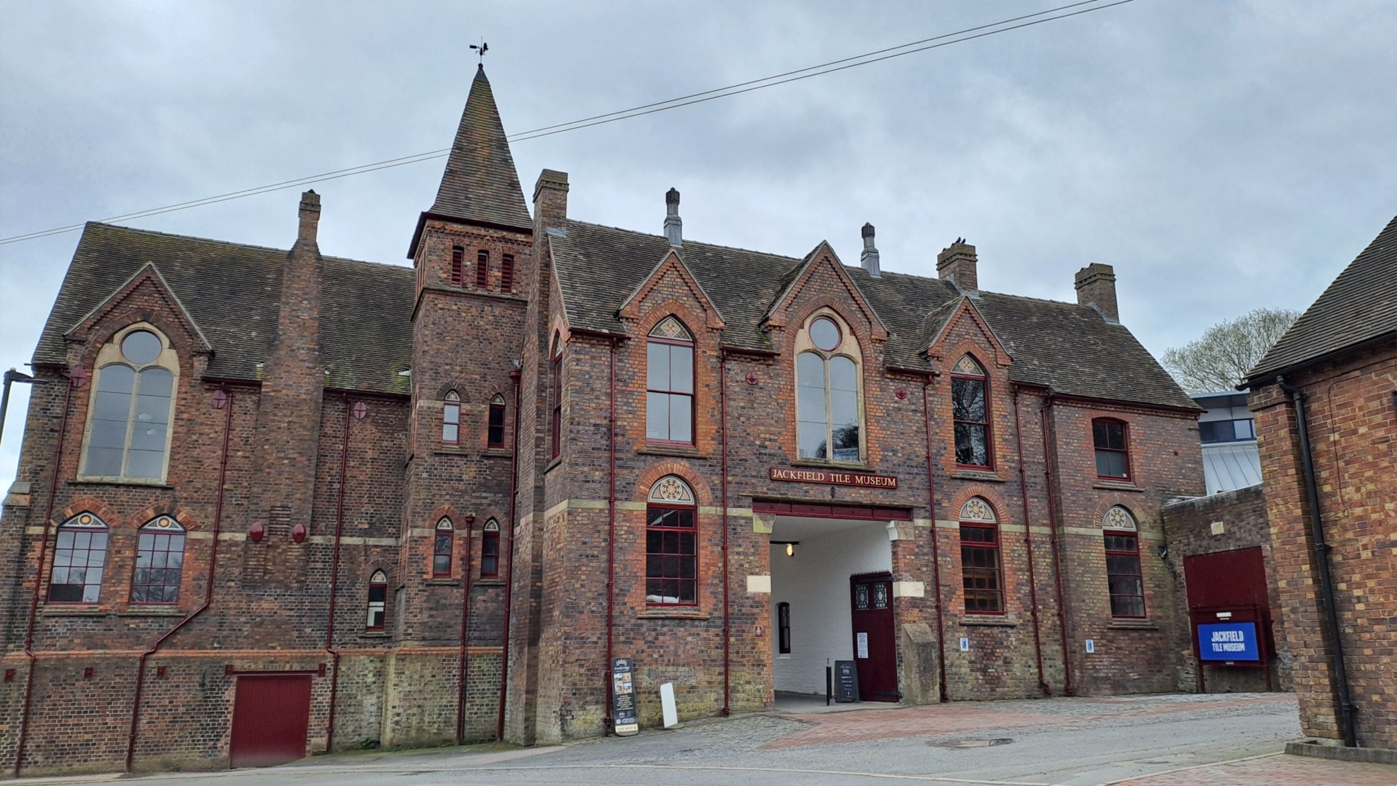 Red-brick Victorian-style building with pitched roofs, chimneys and a central tower, viewed from the road at the front, under an overcast sky. A sign above the entrance reads 'Jackfield Tile Museum,' with tall arched windows and decorative brickwork visible across the façade.