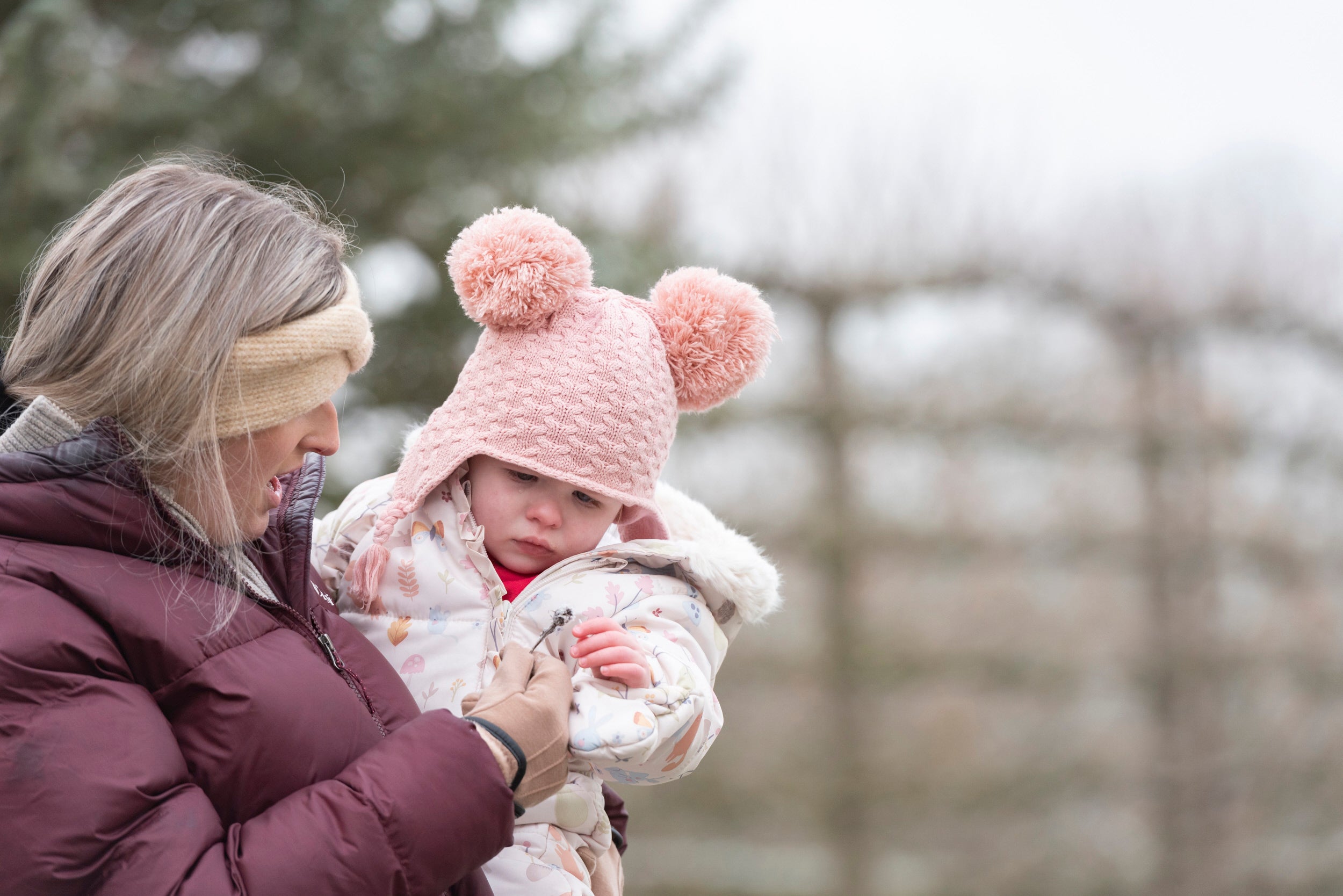 Woman carrying young child in a Garden, both wearing hats and gloves.
