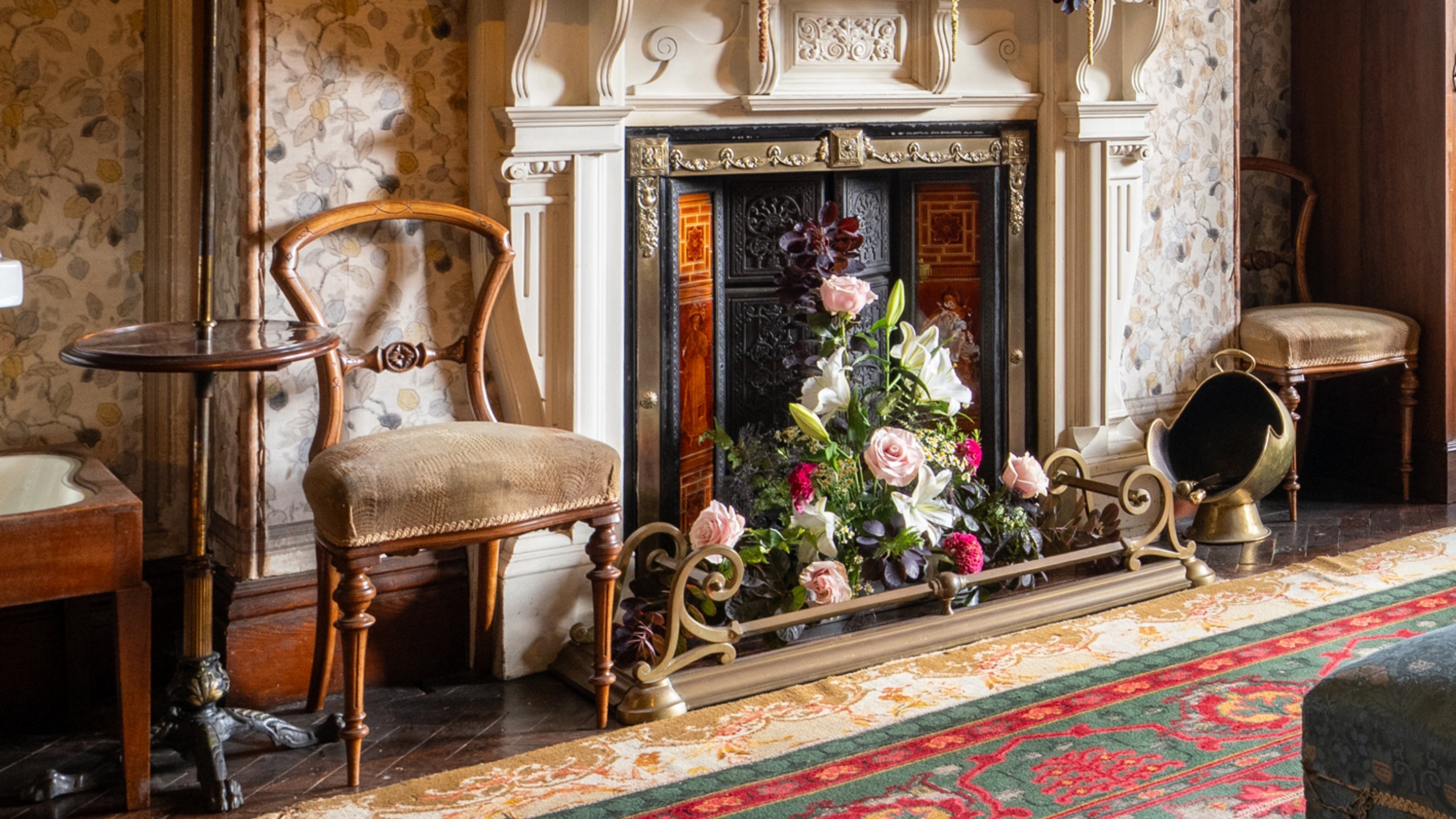 Vintage fireplace dressed with a floral arrangement of roses, lilies and greenery, framed by ornate tiles and surrounded by period furniture in a historic Sunnycroft interior.