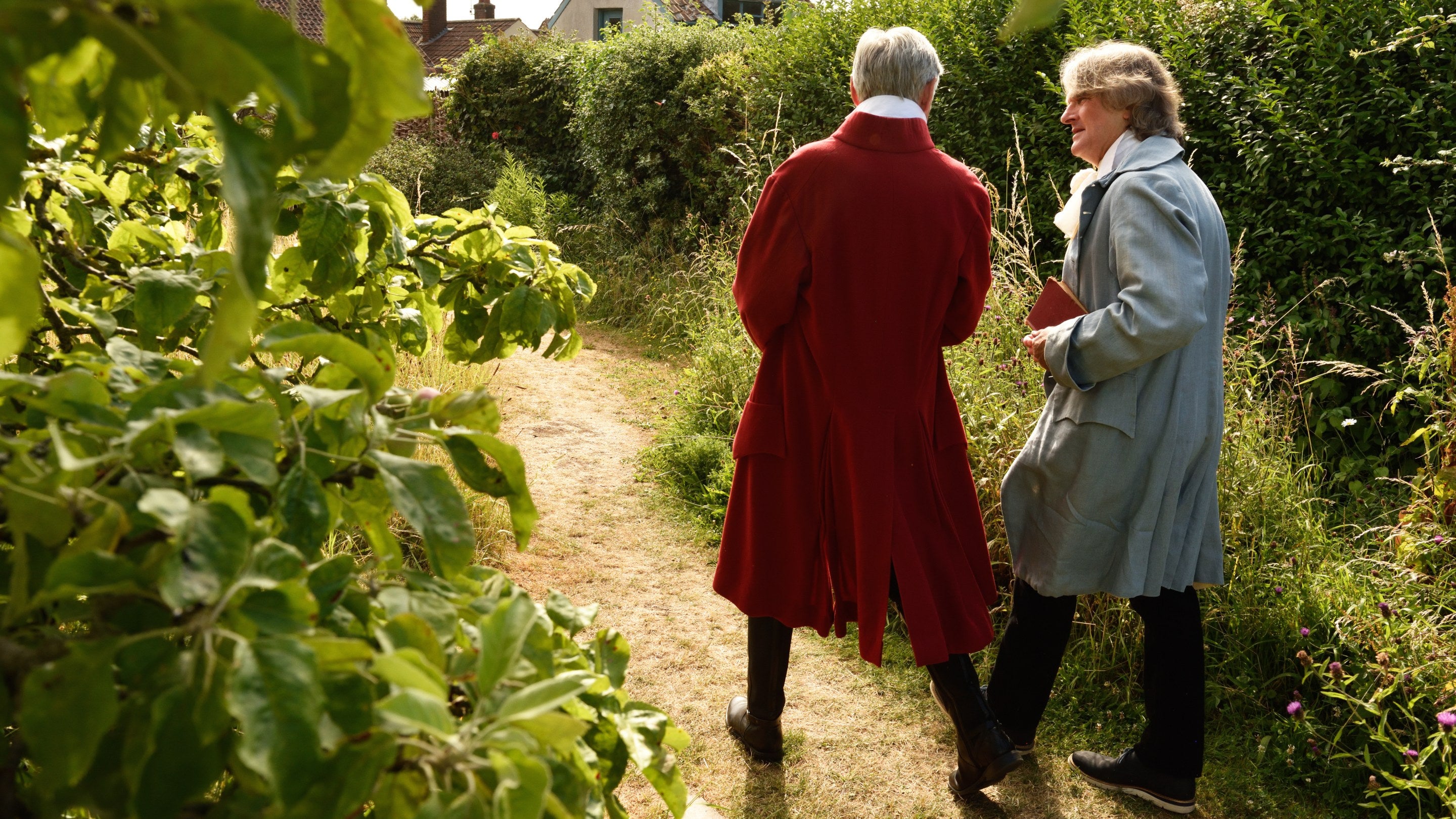 Two volunteer interpreters in 18th-century costumes walk down a path in the garden at Coleridge Cottage, Somerset