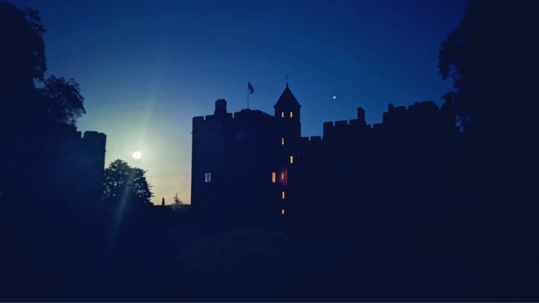 Dunster Castle silhouetted against the night sky. The windows of a tower on the left are illuminated with orange light and there is a bright moon to the left