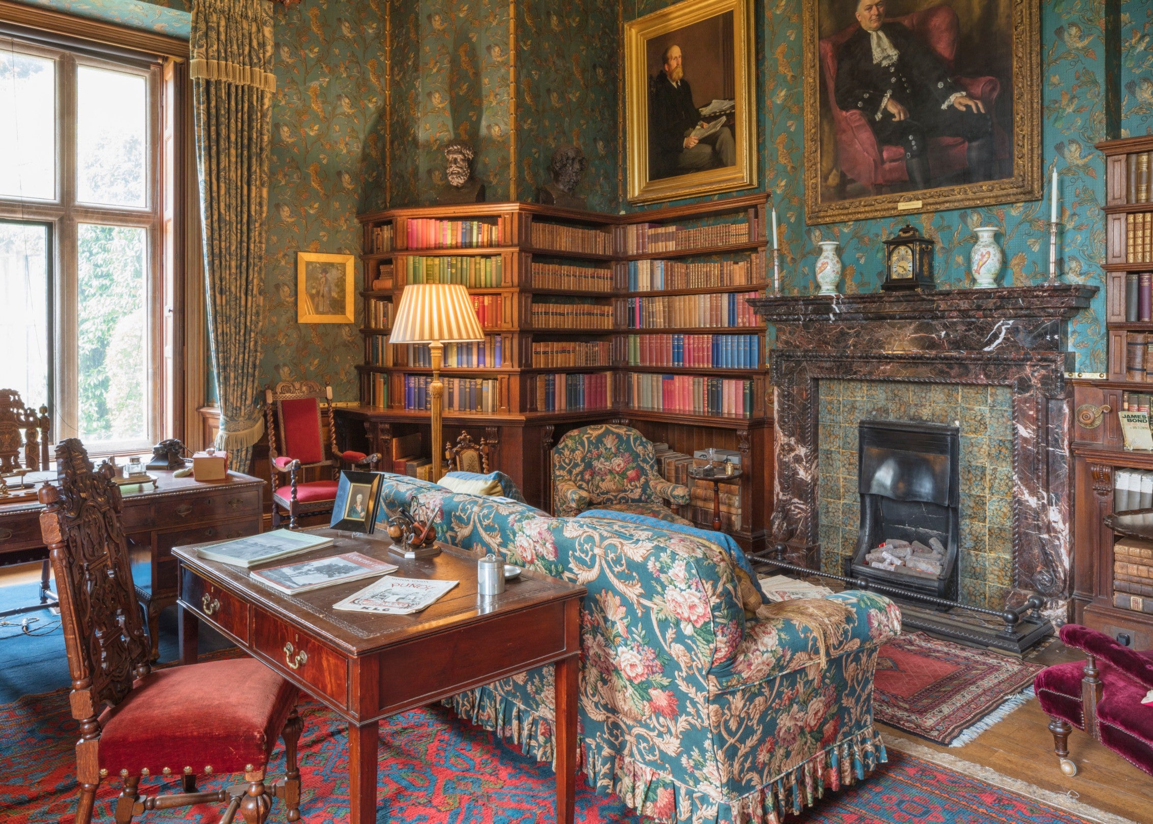 A library with ornate blue wallpaper, a marble fireplace in the centre, and dark wood bookshelves. There are a blue sofa and armchair and wooden desk and chair in the room.