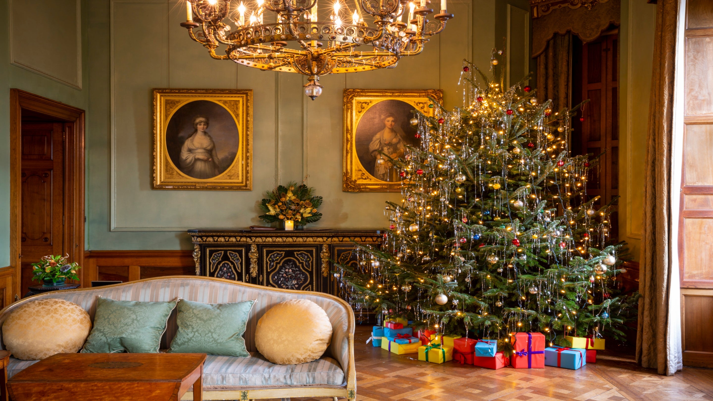The drawing room at Dunster Castle decorated for Christmas. A large, beautifully lit Christmas tree stands to the right, adorned with golden lights and ornaments, with colourful wrapped presents underneath. Two framed portraits hang on the wall above an ornate black and gold sideboard decorated with festive greenery. A chandelier with warm lights hangs from the ceiling, and a pale blue sofa with round cushions sits in the foreground on a polished wooden floor.