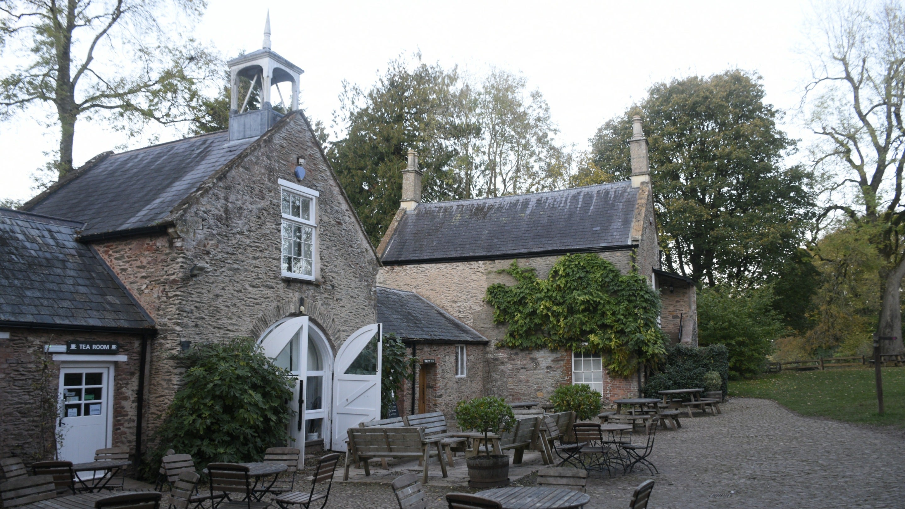 View of an open cobbled courtyard, with a range of historic buildings on one side, and wooden café tables and chairs
