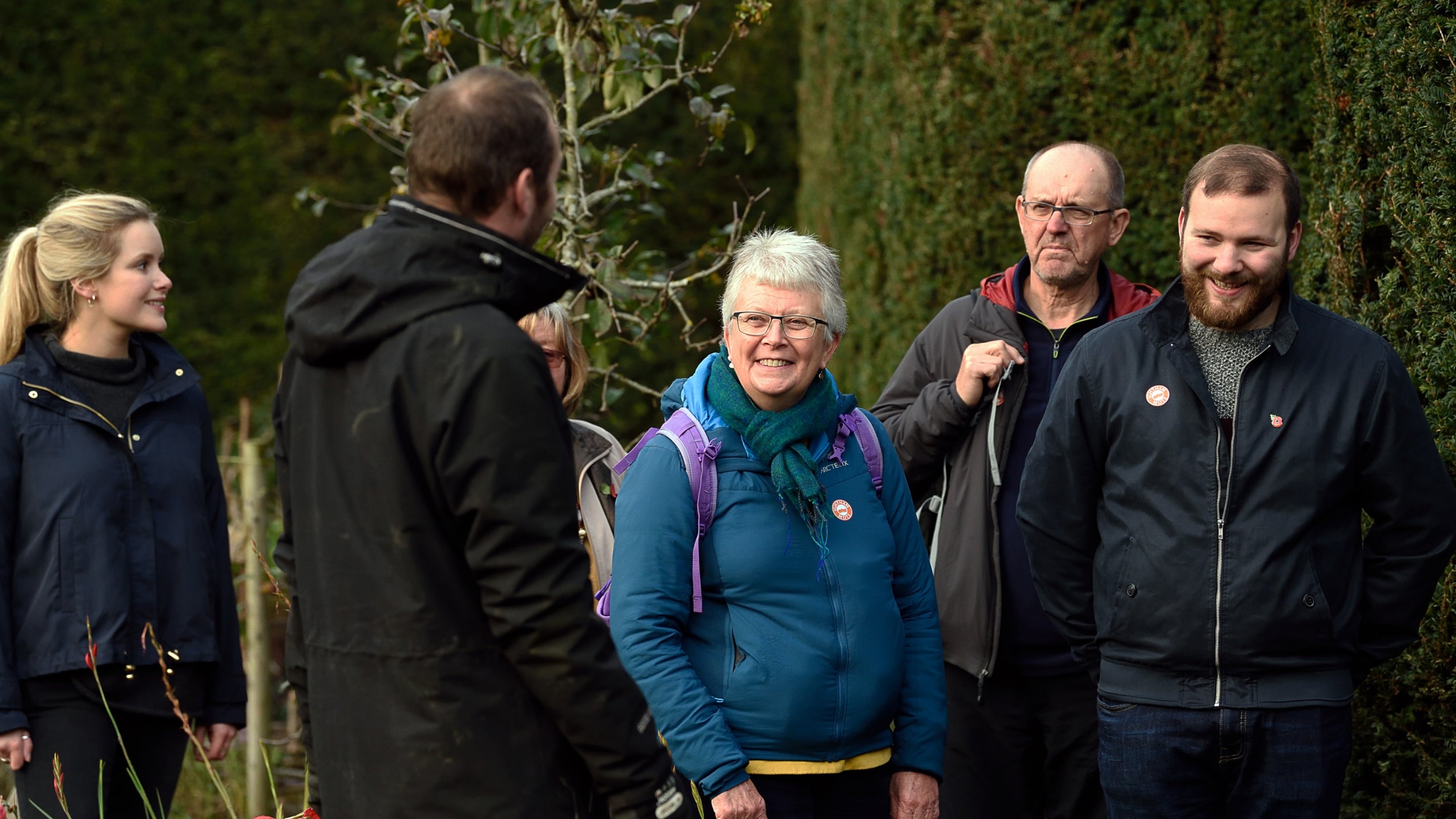 A group of people in front of a yew hedge, smiling as they listen to a guide.