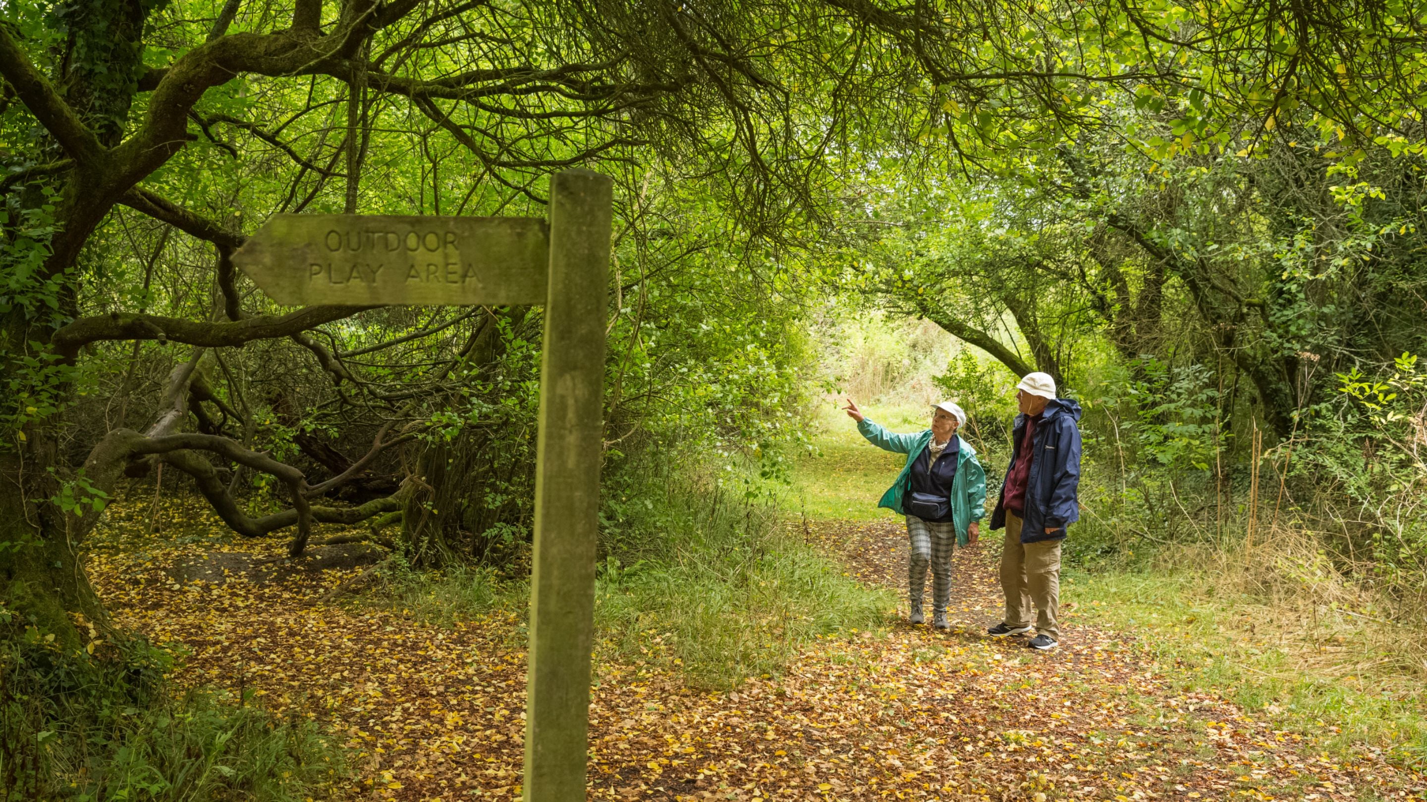 Two walkers on a woodland path covered in fallen leaves look at a signpost that reads 'Outdoor Play Area'