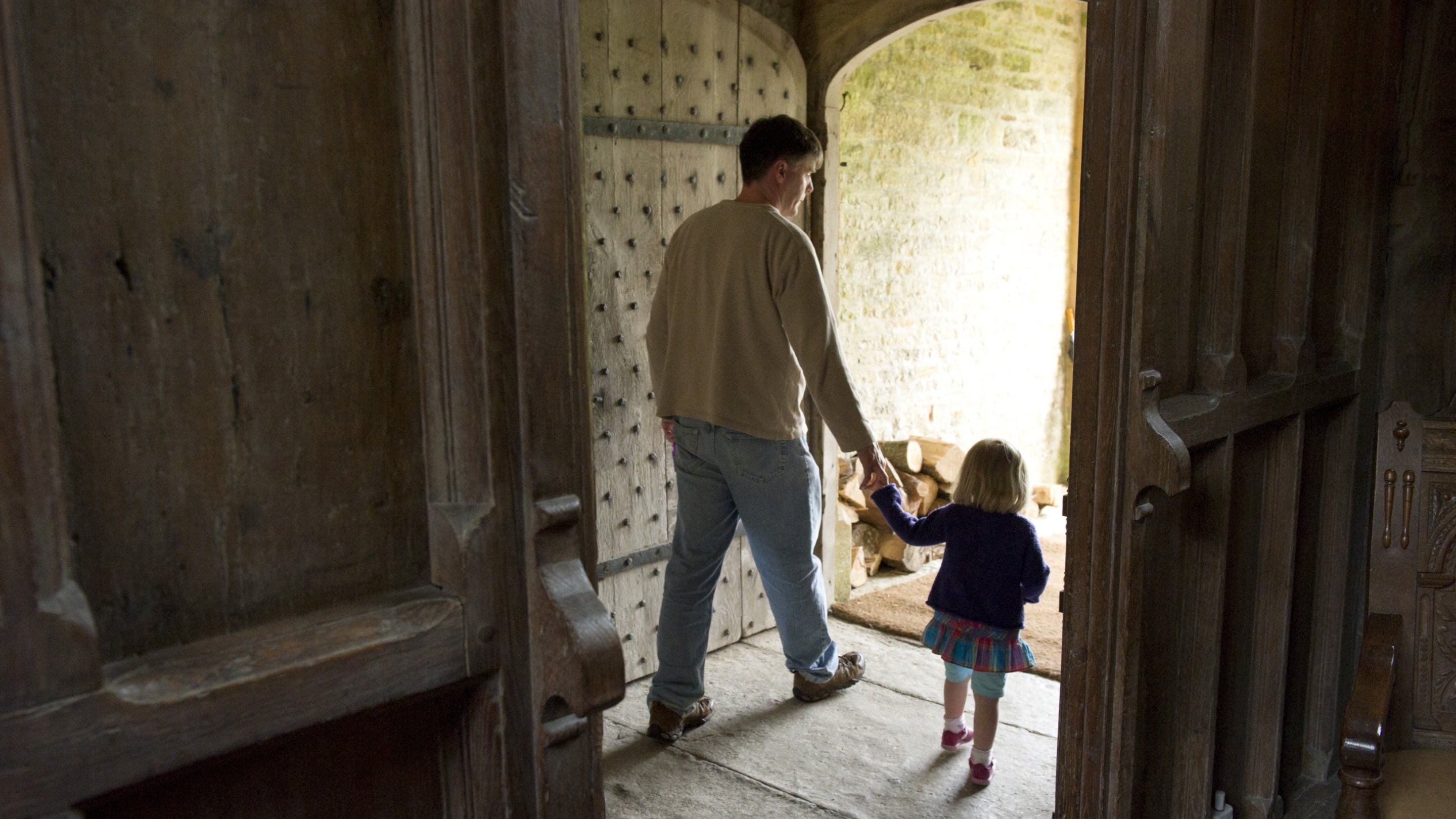 A father and daughter hold hands and walk out over flagstones through an old wooden doorway