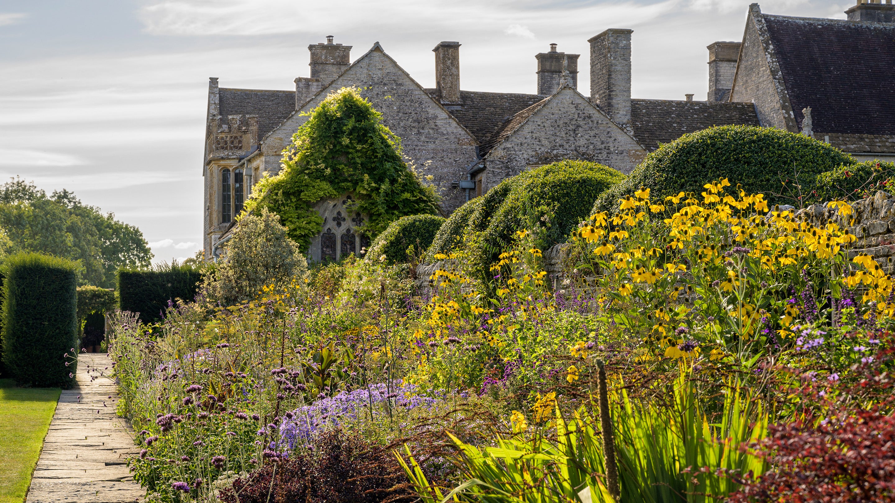 A border of plants and flowers in bloom with a view of Lytes Cary Manor behind