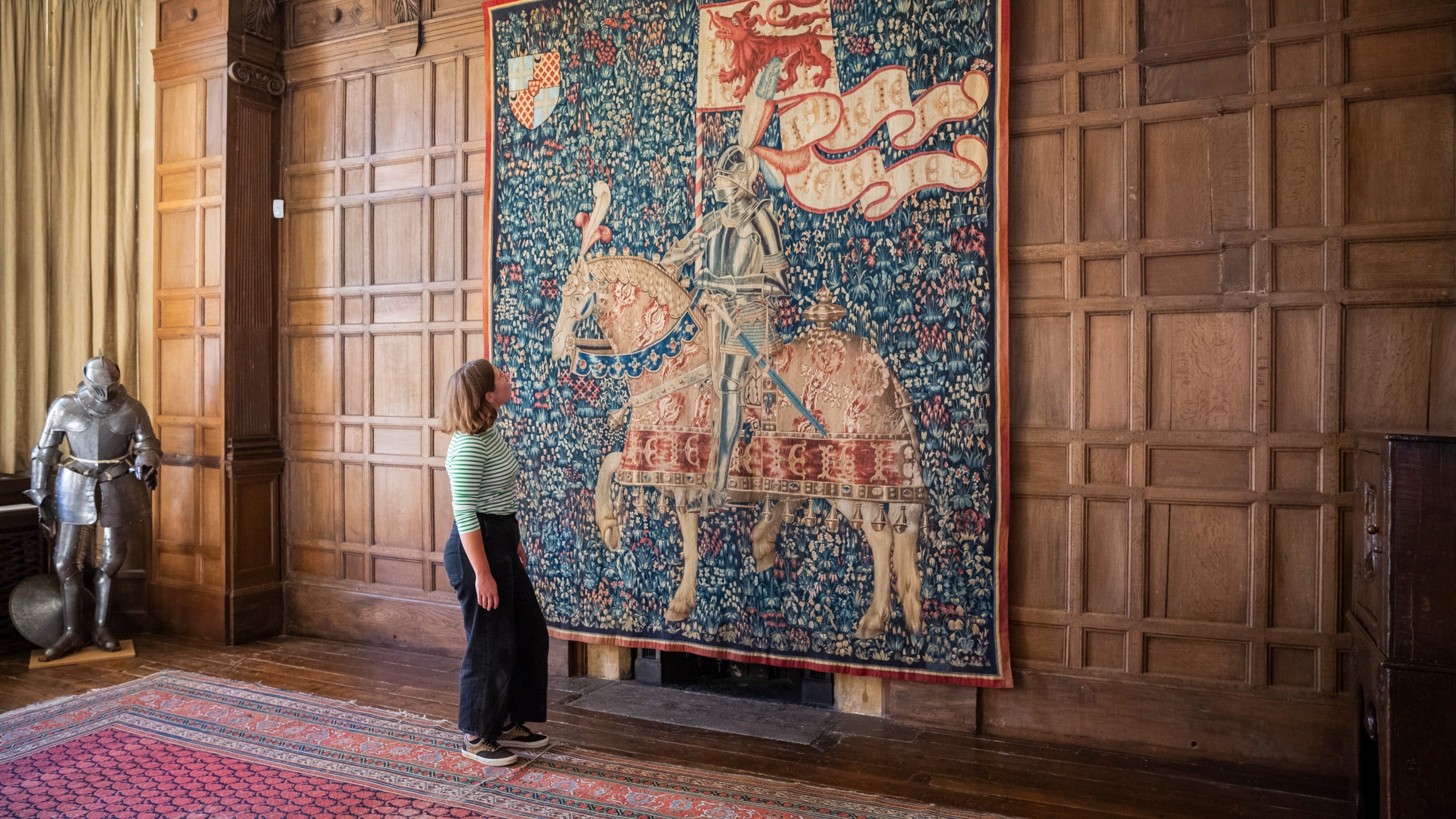Young woman in grand wood panelled gallery looking at tapestry of a knight