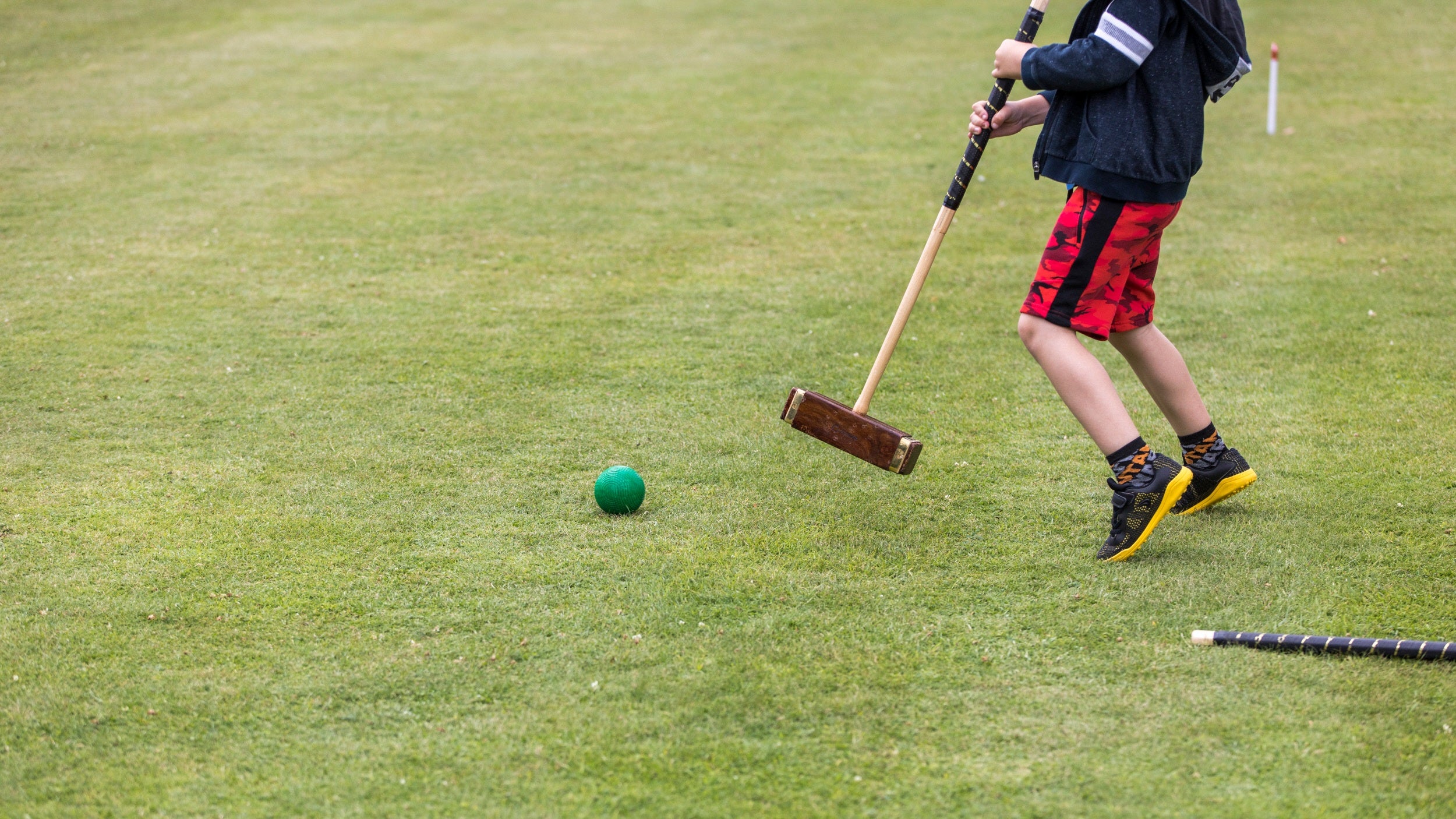 The torso and legs of a boy holding a croquet mallet on a closely cropped mown lawn.