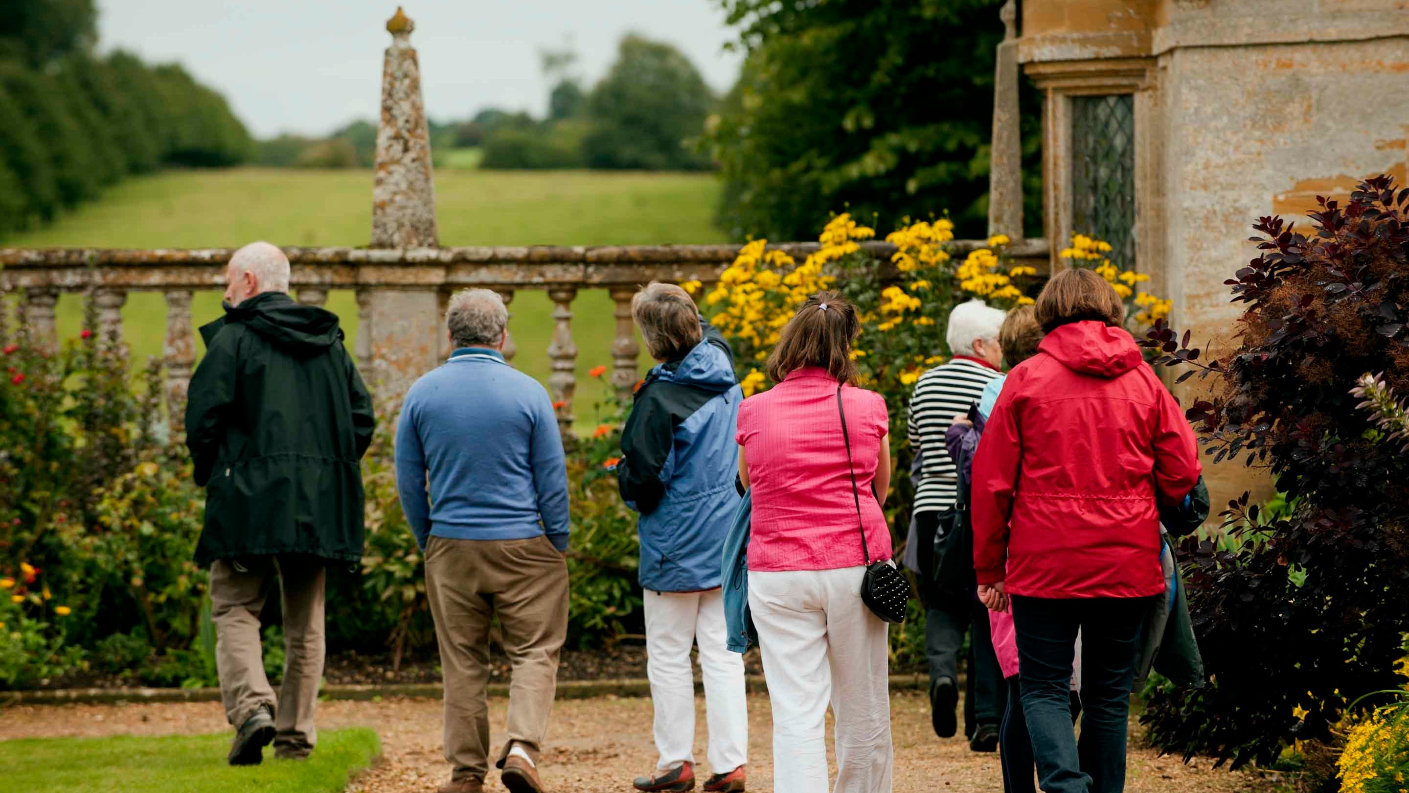 Seven visitors walking away from the camera as they explore the east court garden at Montacute, Somerset. Borders of shrubs line the path as they walk towards a stone balustrade and pavilion with the parkland stretching away into the distance.