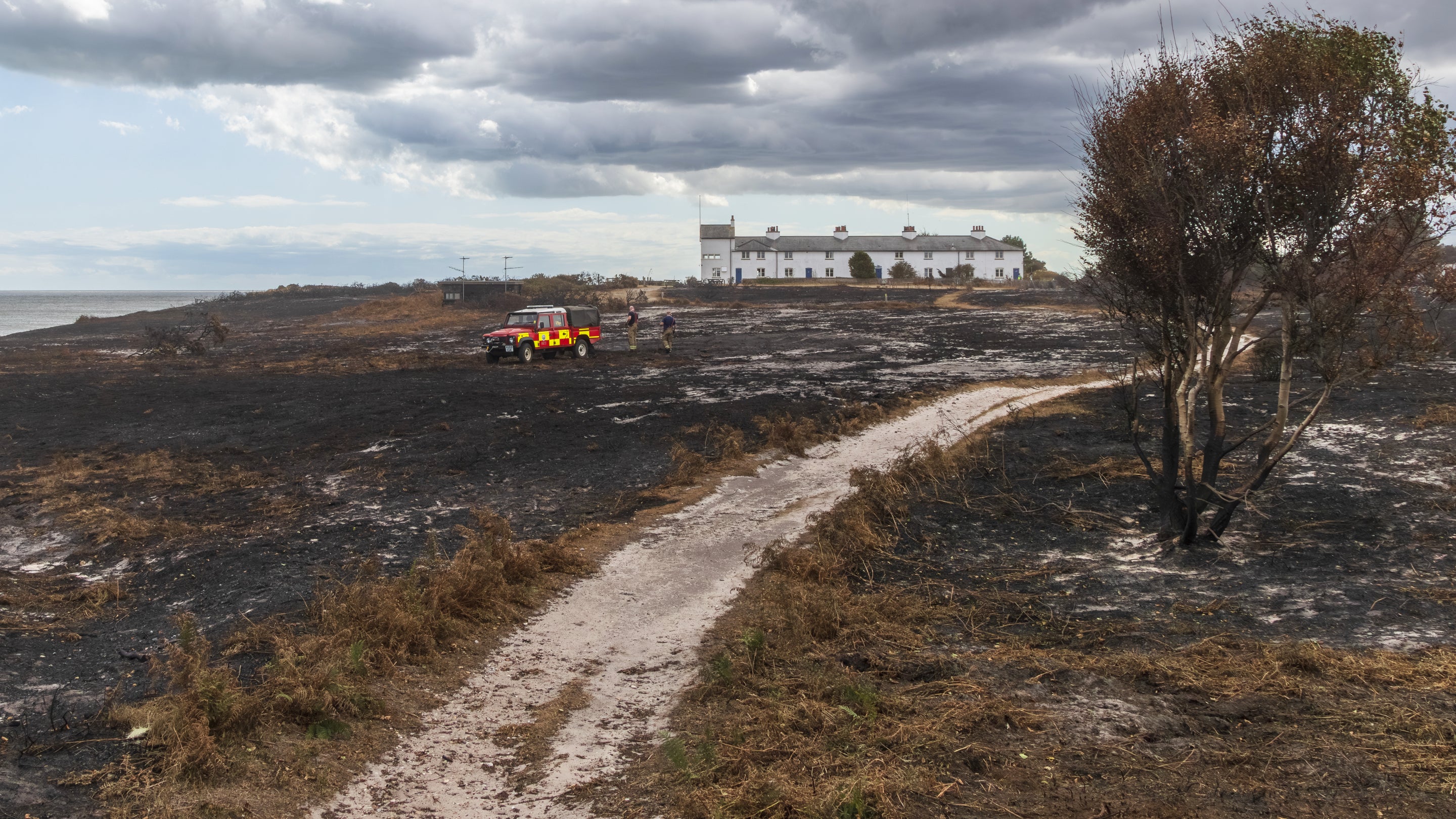 A fire service vehicle surveys burnt heathland at Dunwich Heath following a wildfire, with dark storm clouds overhead and buildings in the distance.