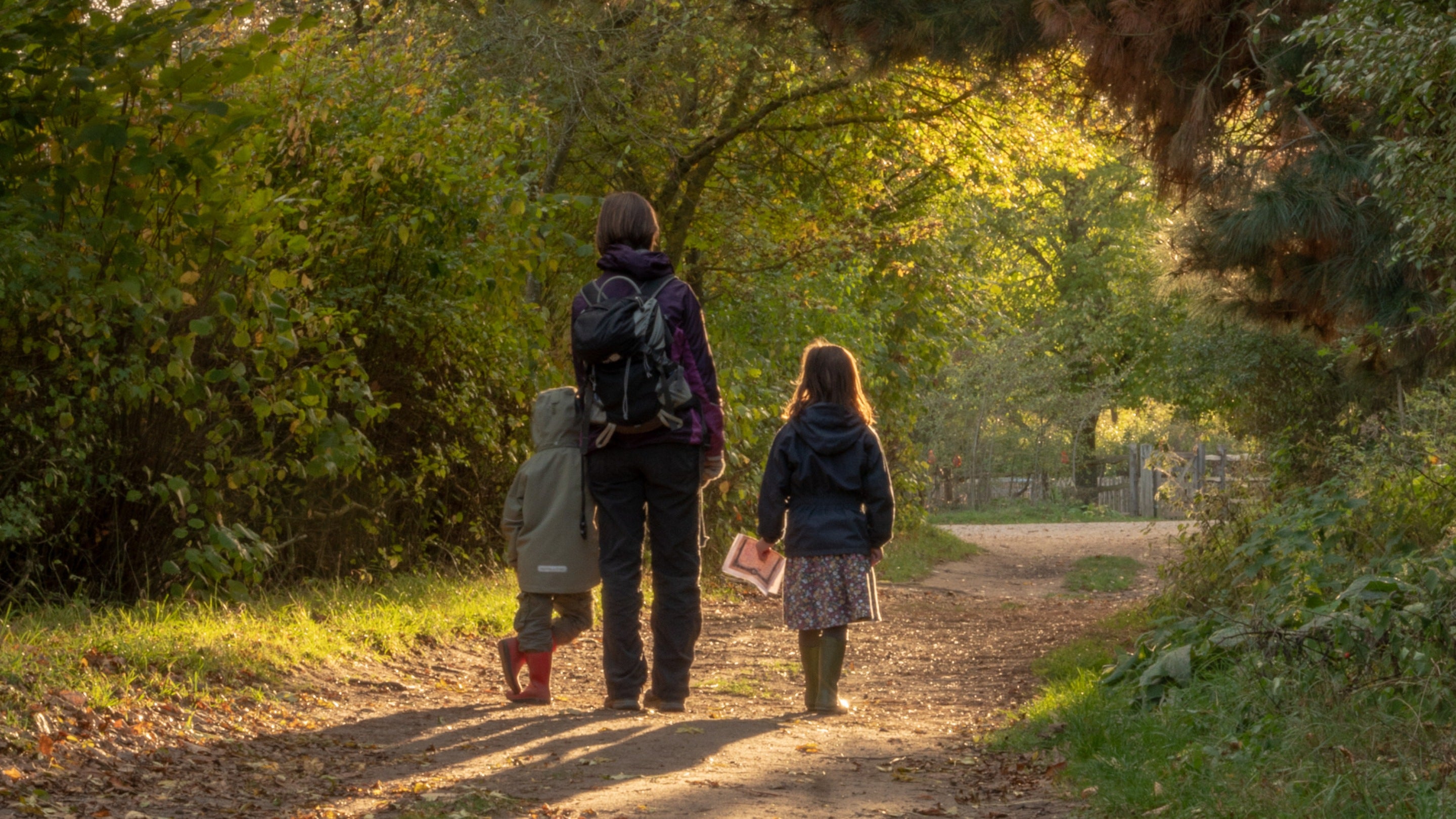 A parent and two children wearing winter coats walk through autumn trees in golden late afternoon sun