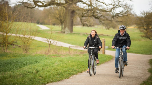 Two visitors cycle side by side through a winding path leading through parkland on the Ickworth Estate. They're wrapped up in rain coats and the weather appears hazy.