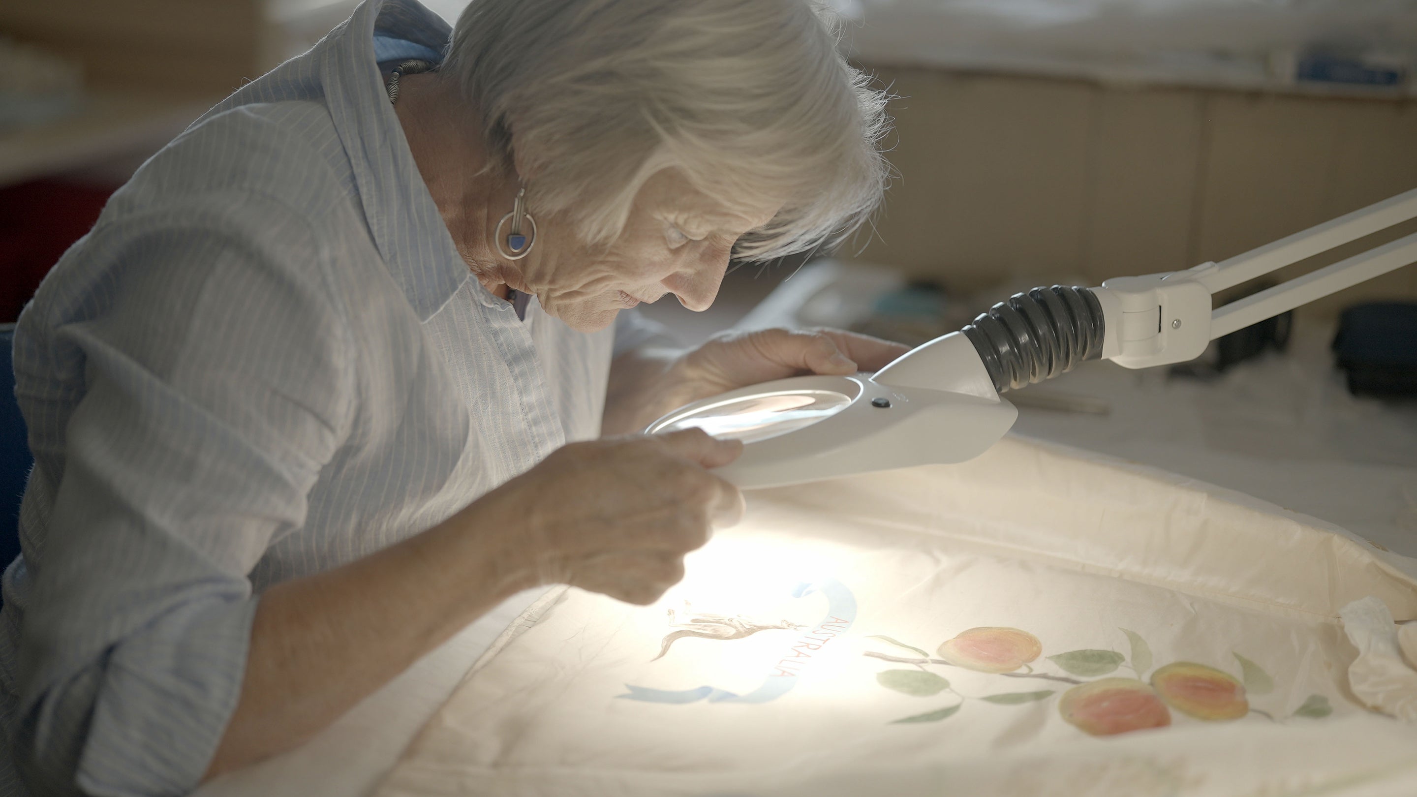 A woman is bent over some white fabric looking through a magnifying glass with a light attached