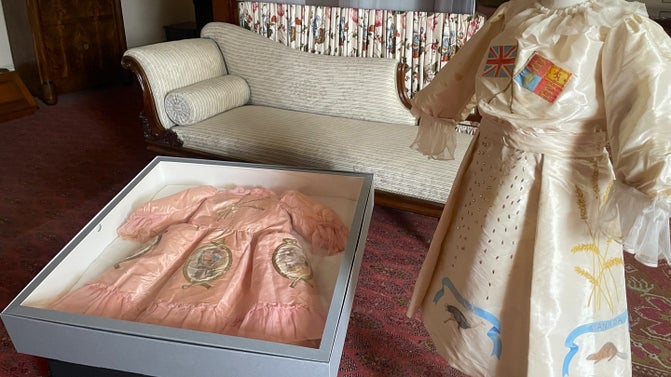 a photo of two dresses with drawings on. One is pink and lain in a box and the other is white and on a stand