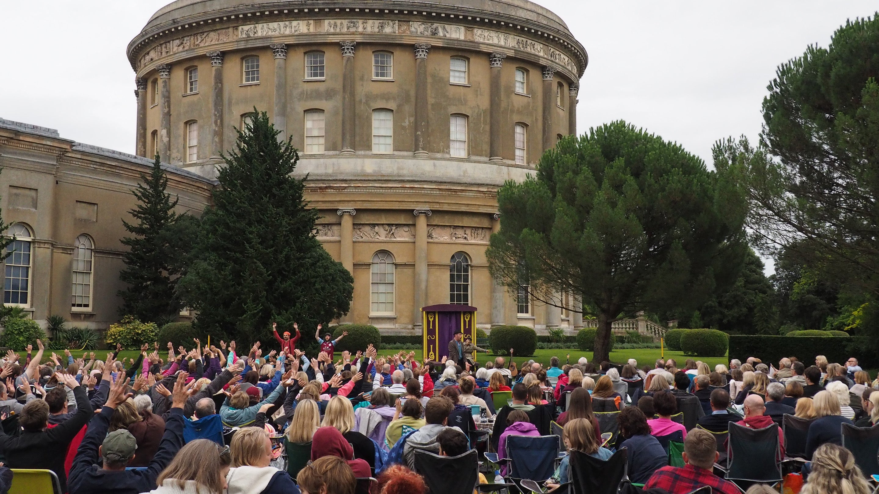 Lots of people sat outside on picnic and camping chairs facing towards an outdoor stage with actors on. the backdrop is the ickworth rotunda and some trees