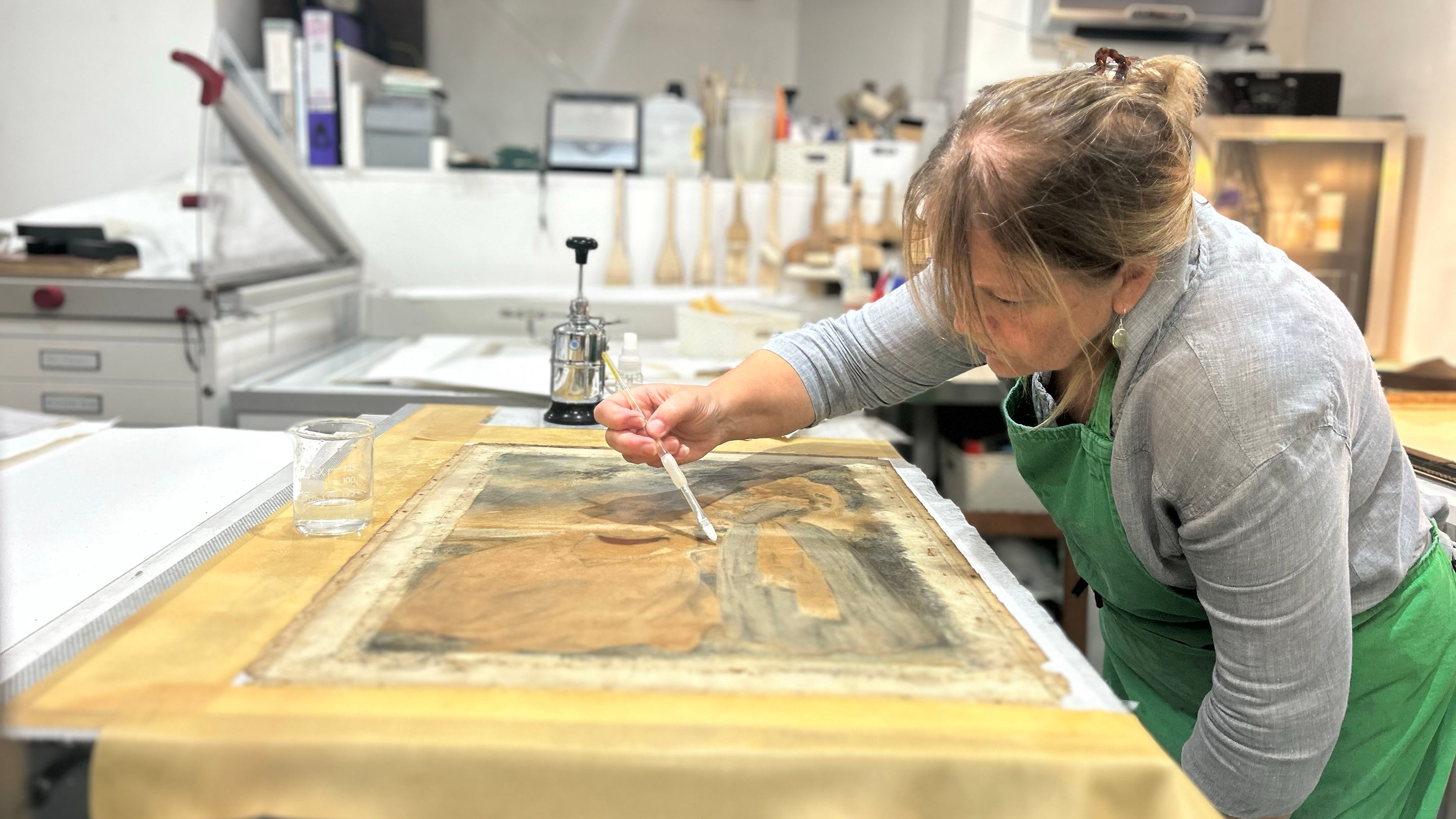 a woman is bent over a painting that is lain flat on a table. she has a small brush that she is carefully sweeping over the surface