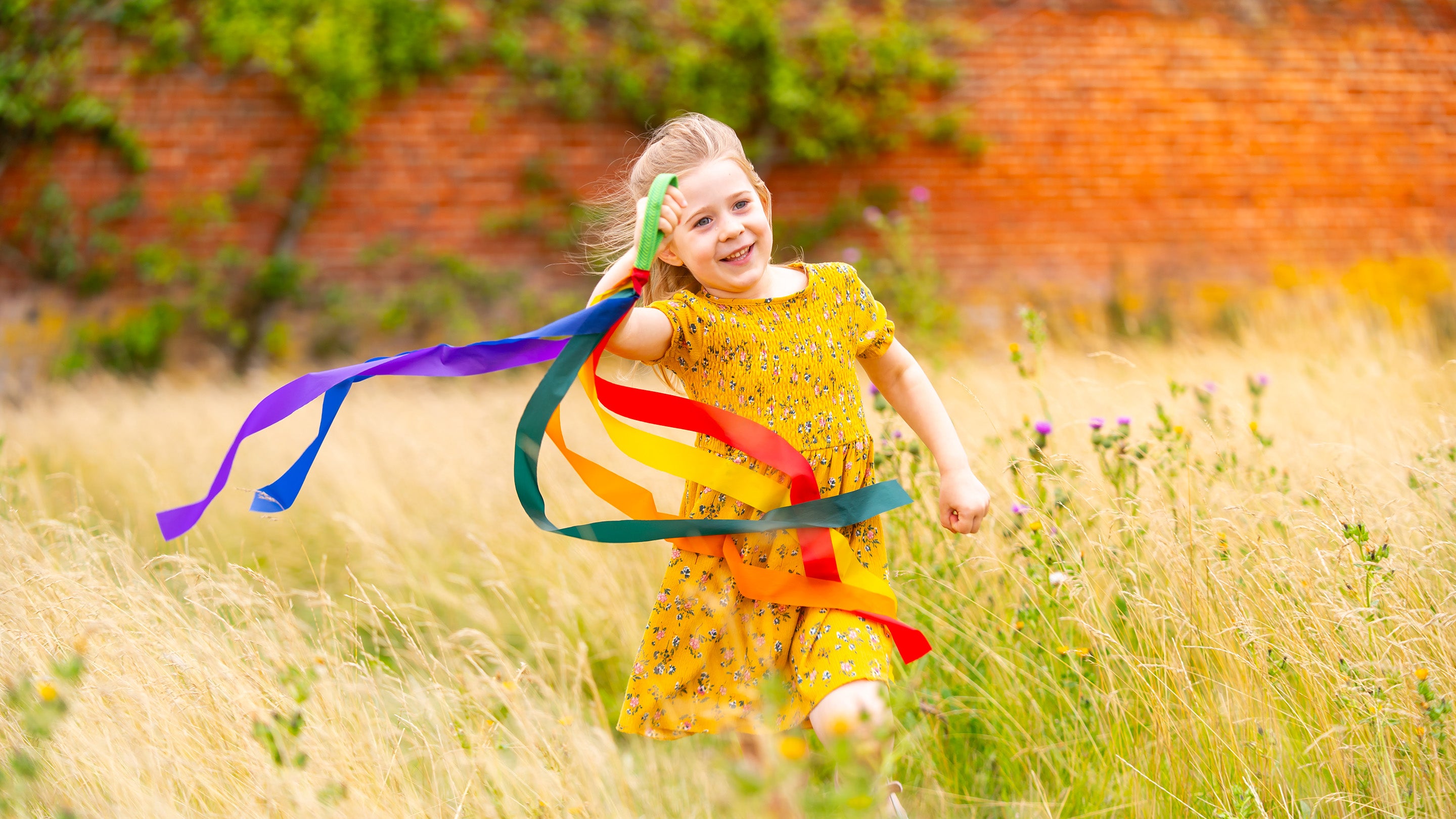 a young girl with blonde hair is running through long grass waving a stick with ribbons attached to it