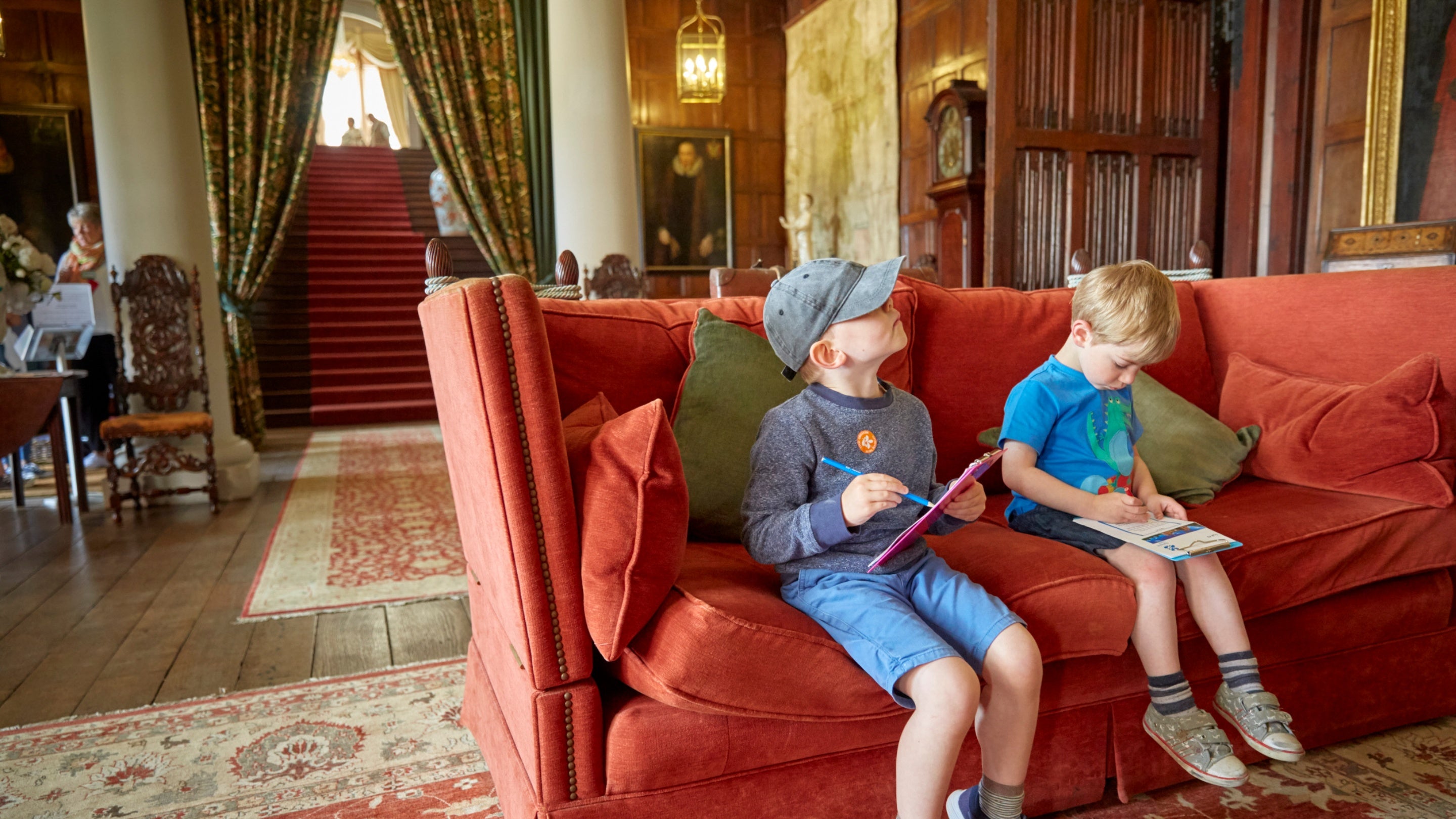 Two children sit on a red sofa in a wood-panelled room, filling out an activity trail