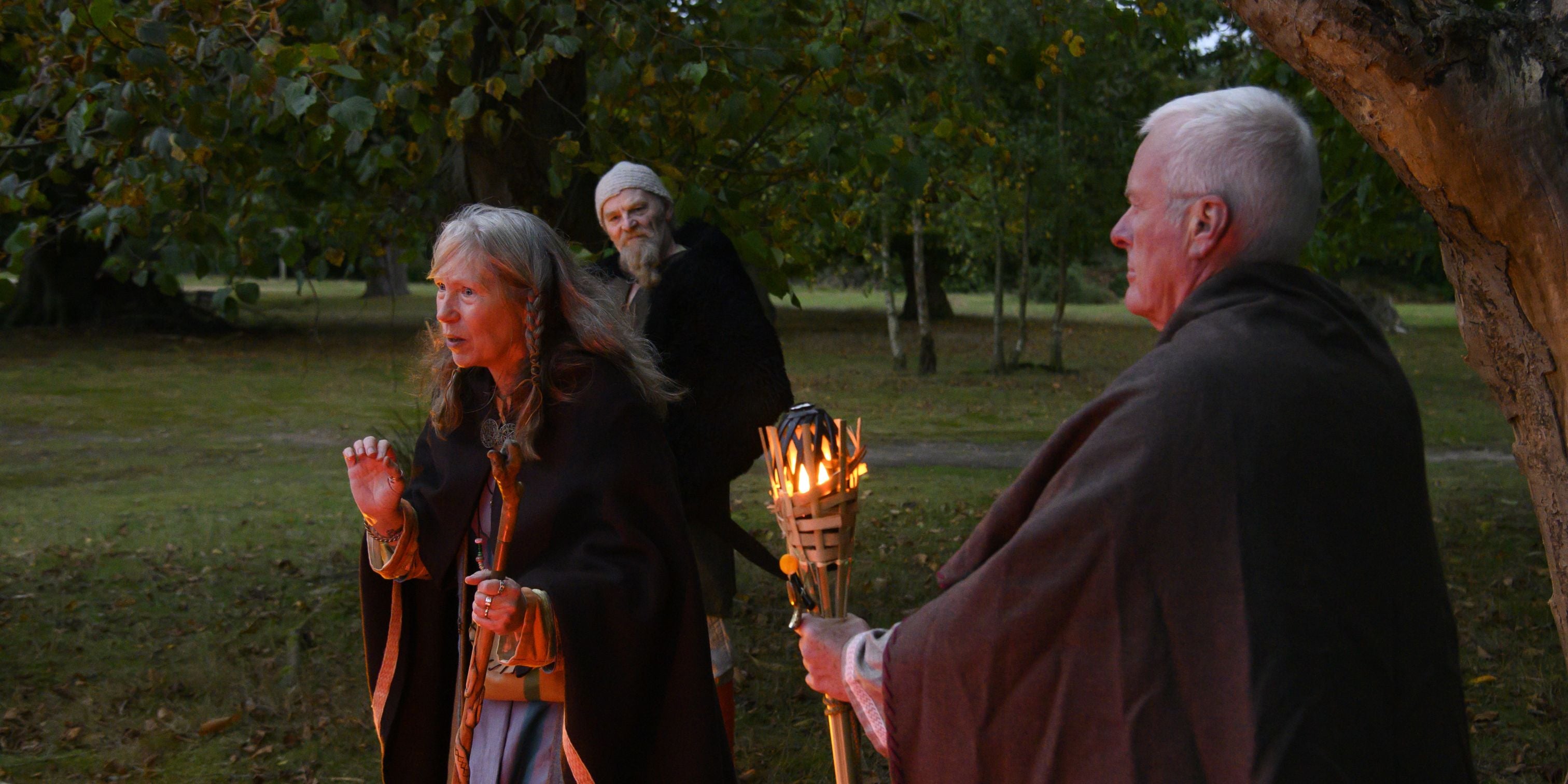 Three people in Anglo-Saxon dress, one holding a lit torch while another is telling a story