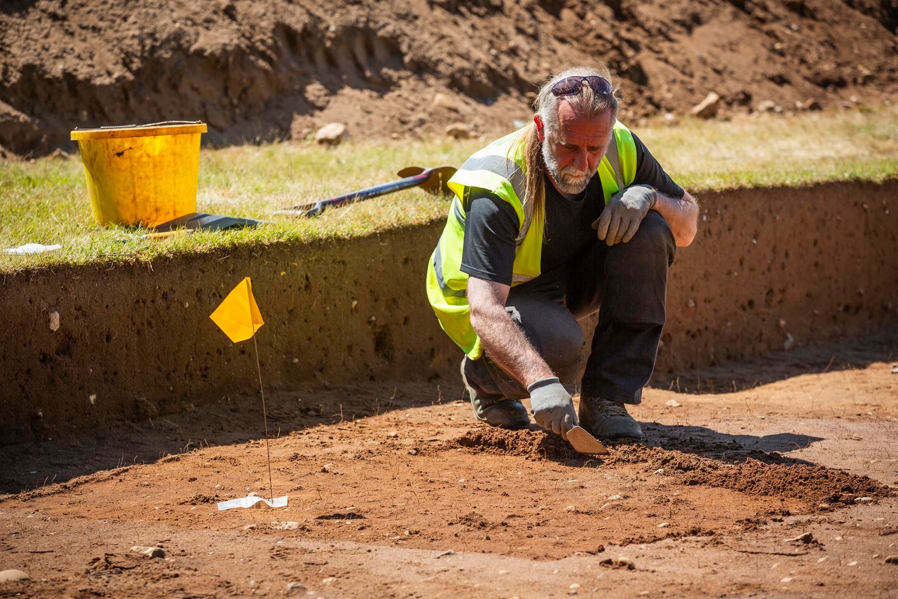 A male archaeologist wearing a hi-vis jacket kneeling in a large trench at Sutton Hoo in Suffolk