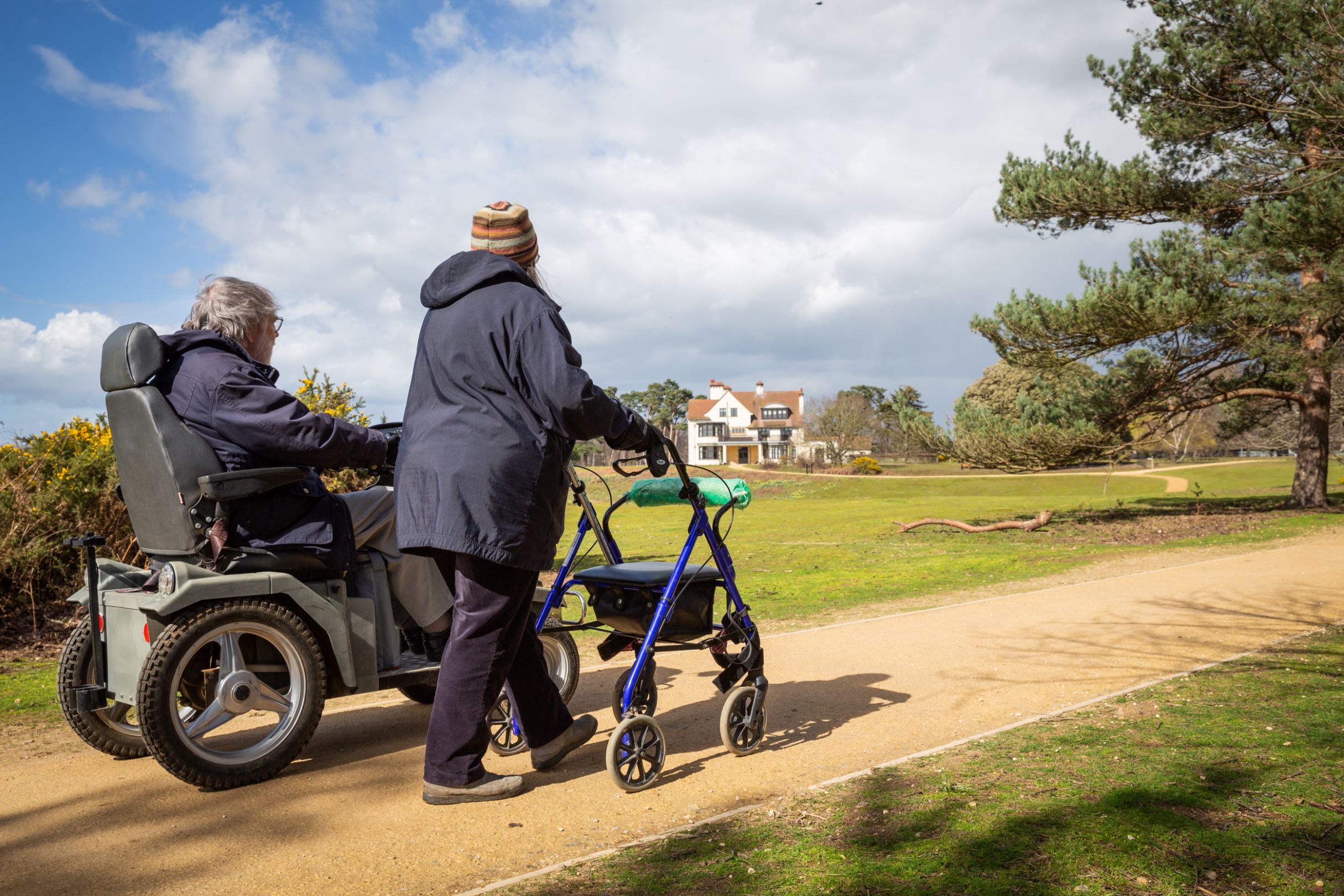 A visitor in a tramper next to a companion with a mobility aid walking towards Tranmer House at Sutton Hoo