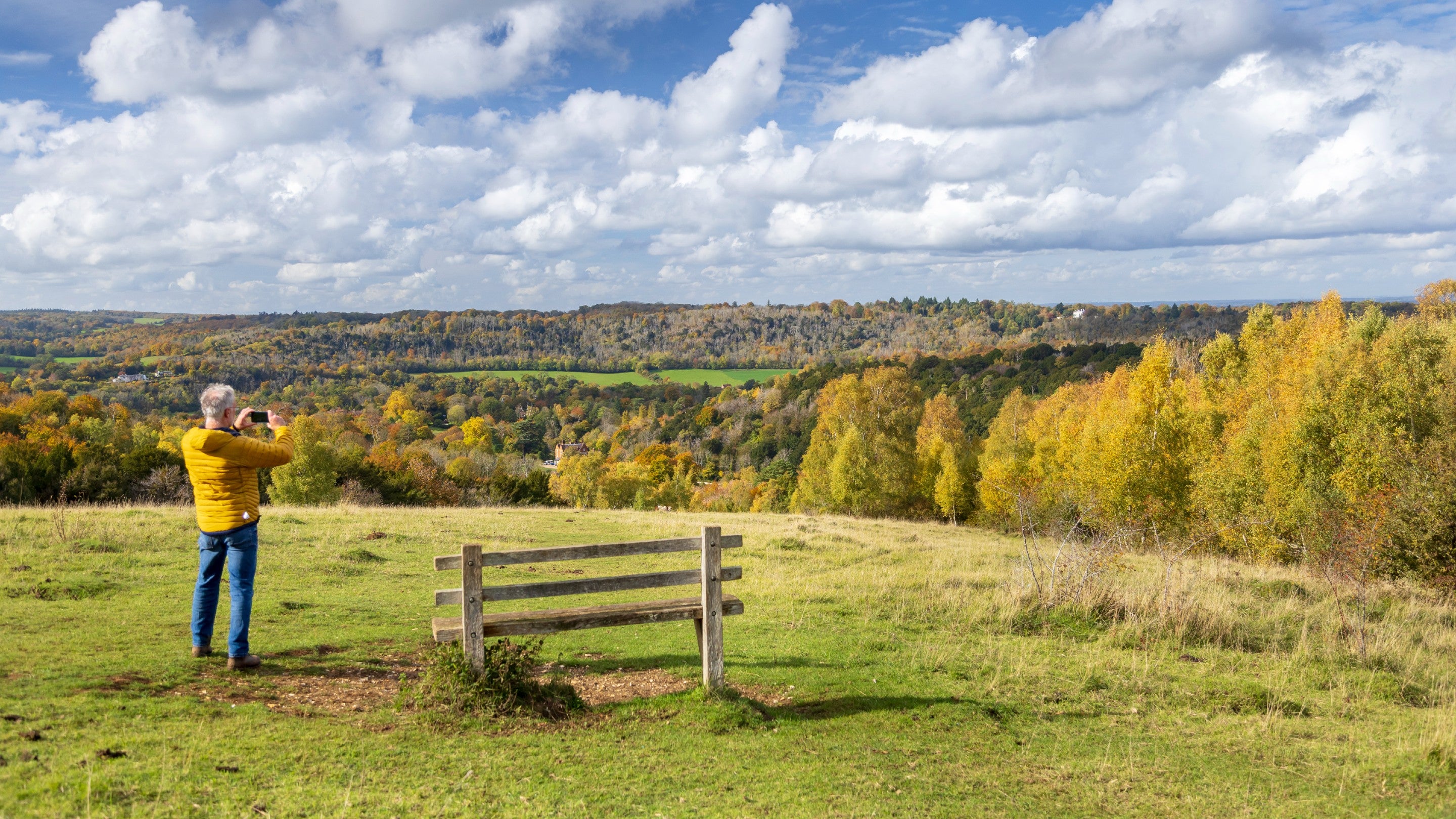 Man in yellow jacket standing by a bench on top of a hill, taking a picture of the view.