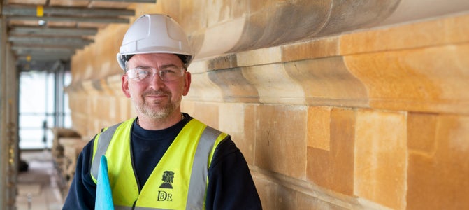 Heritage stonemason Cary Wadey, wearing PPE, stood next to stonework with a blue folder under his arm