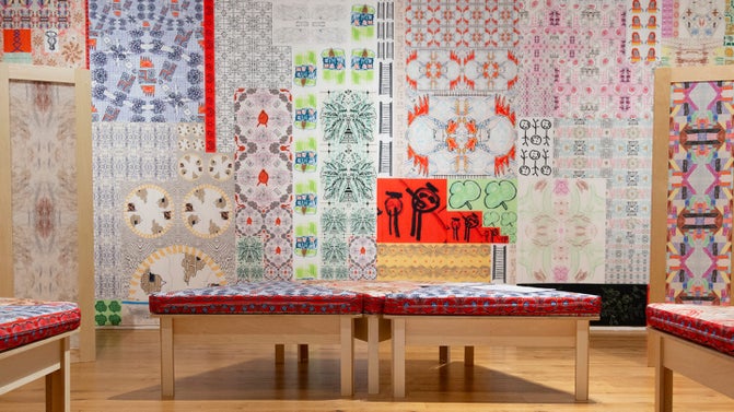 Brightly coloured wallpaper with many patterns on the wall with benches in front of the wall with bright cushions.