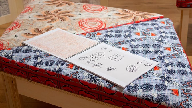 A sketchbook with line drawings is open on a wooden bench with brightly coloured and patterned cushions