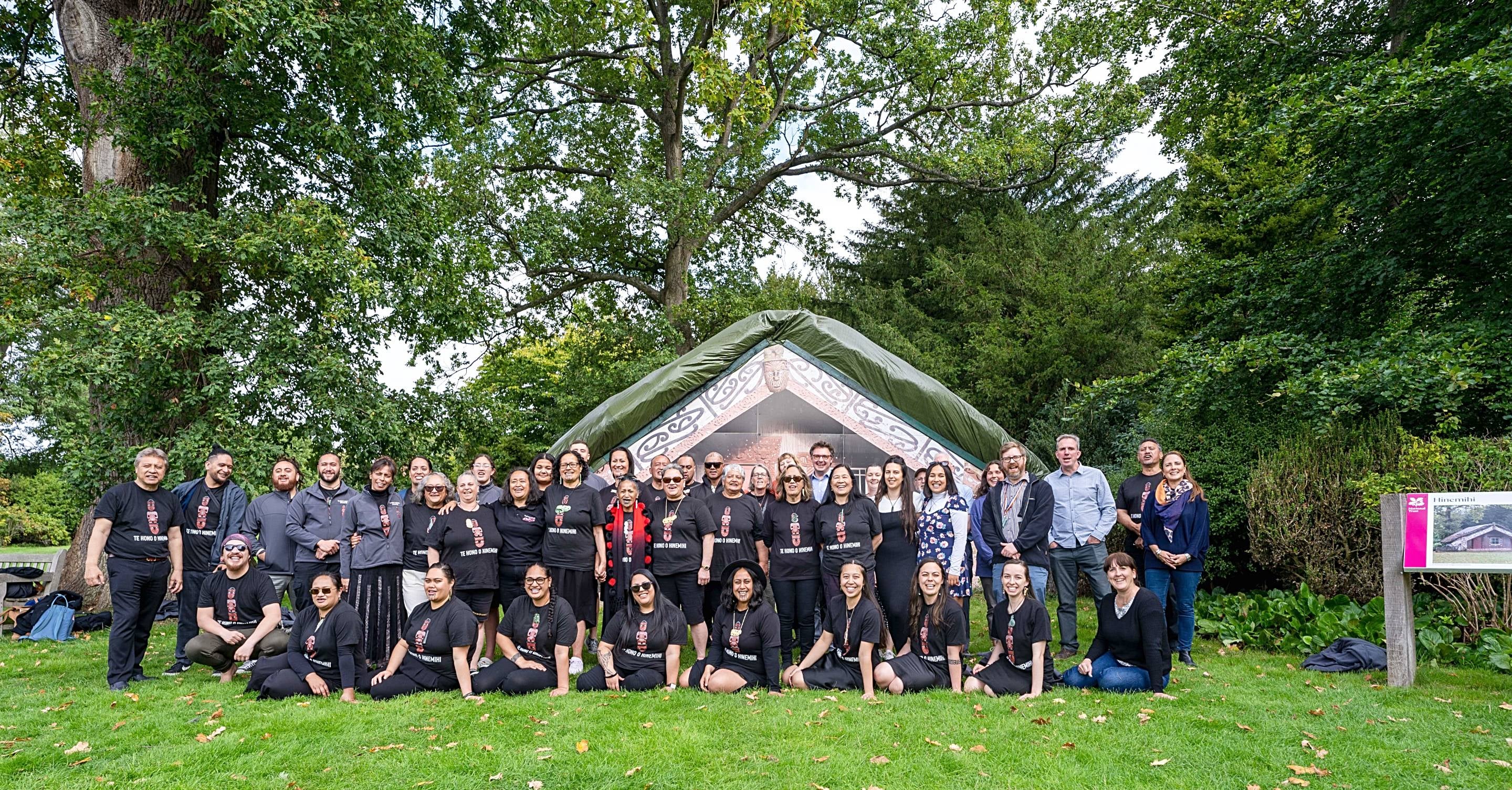 A large group of smiling people gathered in rows on the grass in front of a Maori meeting house surrounded by trees